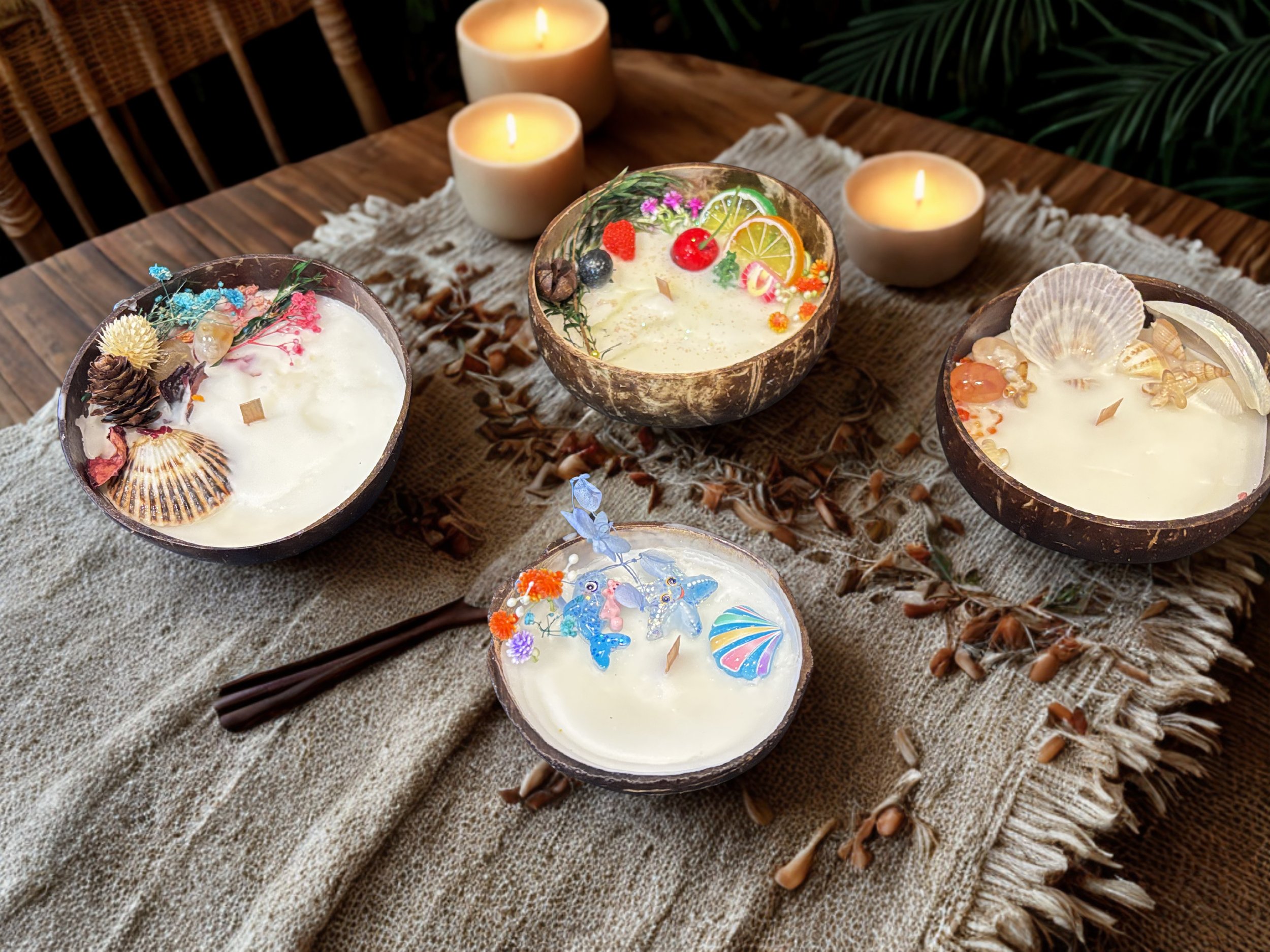Decorative bowls filled with creamy desserts topped with colorful candies, flowers, chocolate, seashells, and other decorations on a textured cloth, with lit candles and green foliage in the background.