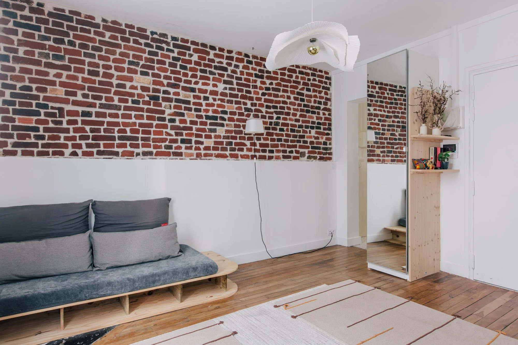 Intérieur d'un salon avec un mur en briques apparentes, un canapé en bois avec des coussins gris, une lampe murale, un plafond avec suspension en papier blanc et un meuble en bois avec des décorations. Plancher en bois et tapis beige avec rayures