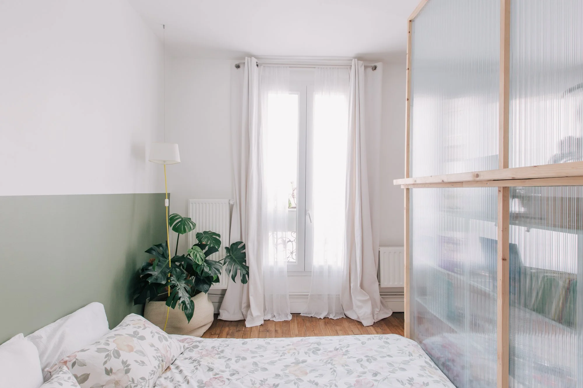 Chambre avec lit à draps floraux, plante verte à côté d'une fenêtre avec rideaux blancs, paroi partielle en panneaux de bois et plastique translucide.