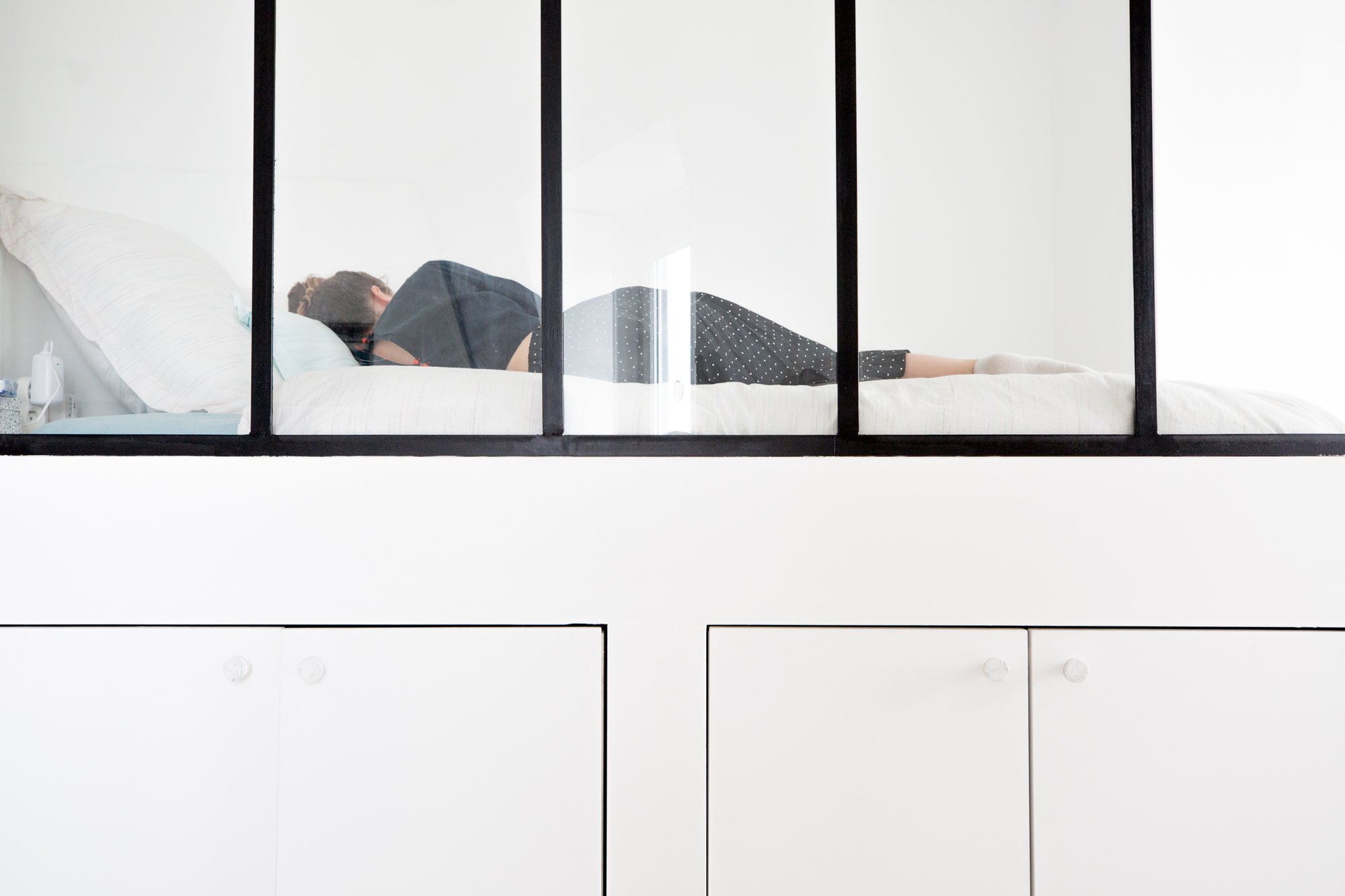 Une femme repose sur un lit dans une pièce blanche, vue à travers une grande fenêtre divisée en plusieurs sections.