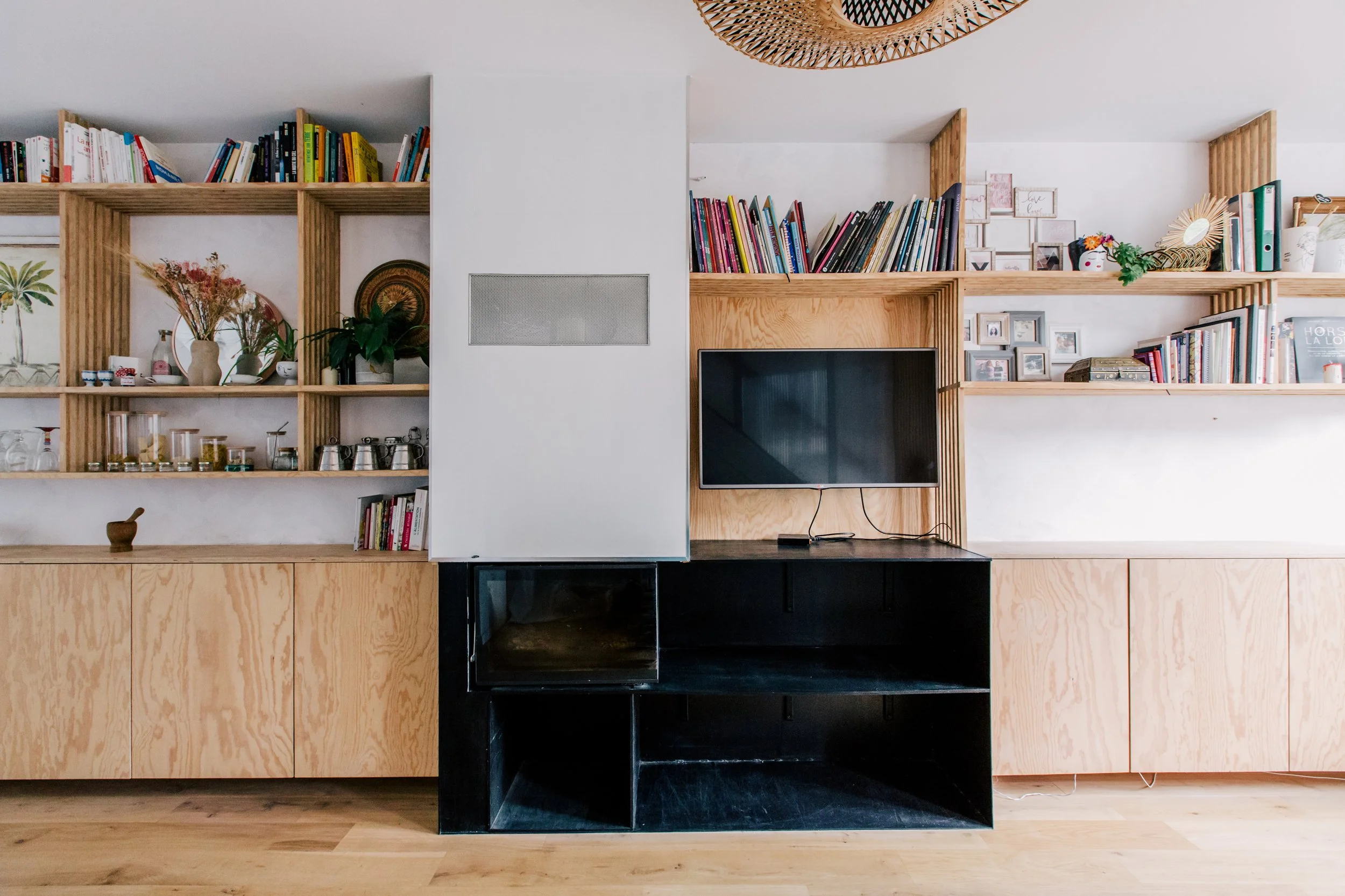 Une étagère en bois contenant des livres, des plantes vertes, des cadres photo et des décorations, avec une télévision murale en dessous, dans un intérieur moderne.