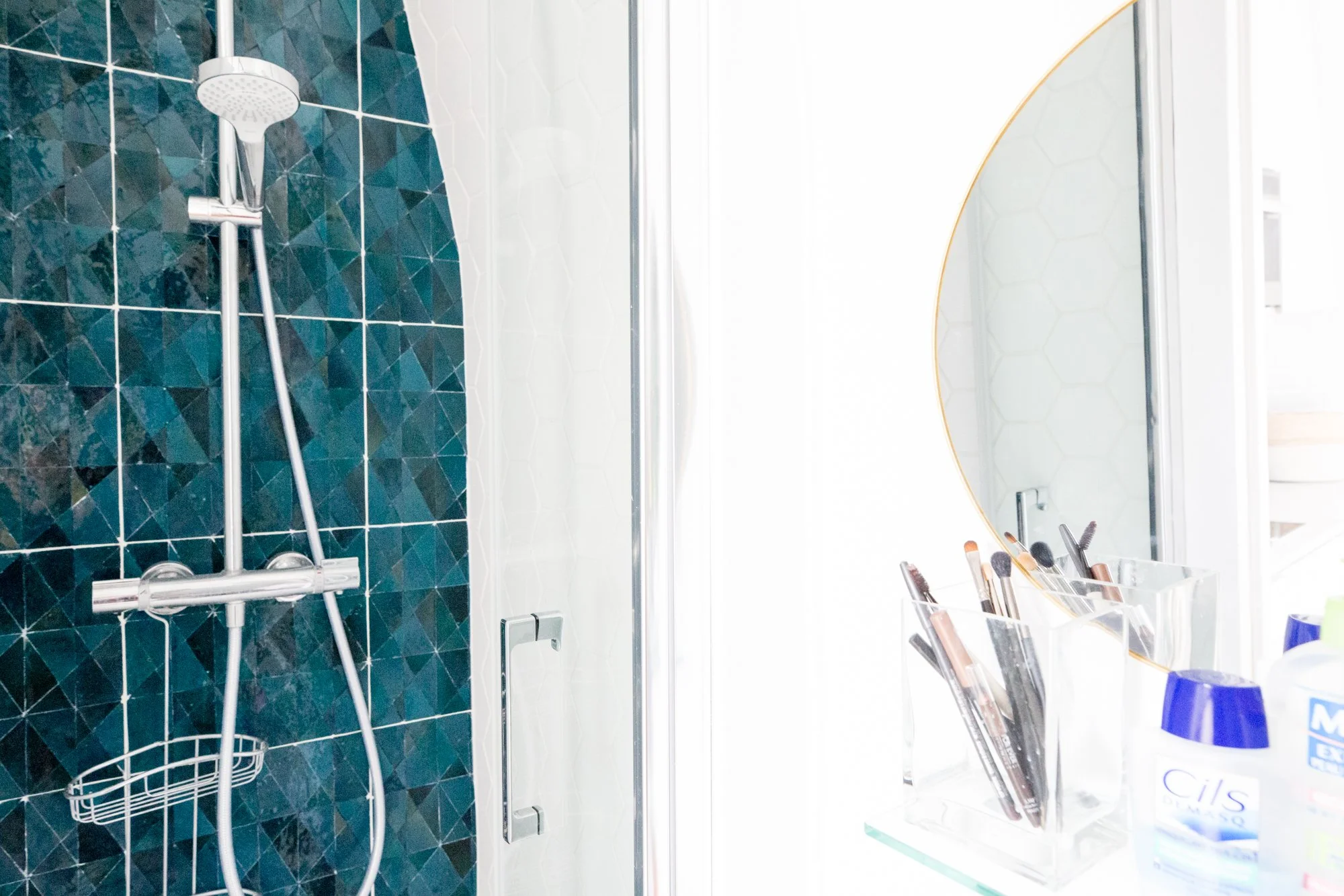 Salle de bain avec douche en verre, carrelage noir et blanc, miroir rond, produits de maquillage sur une table.