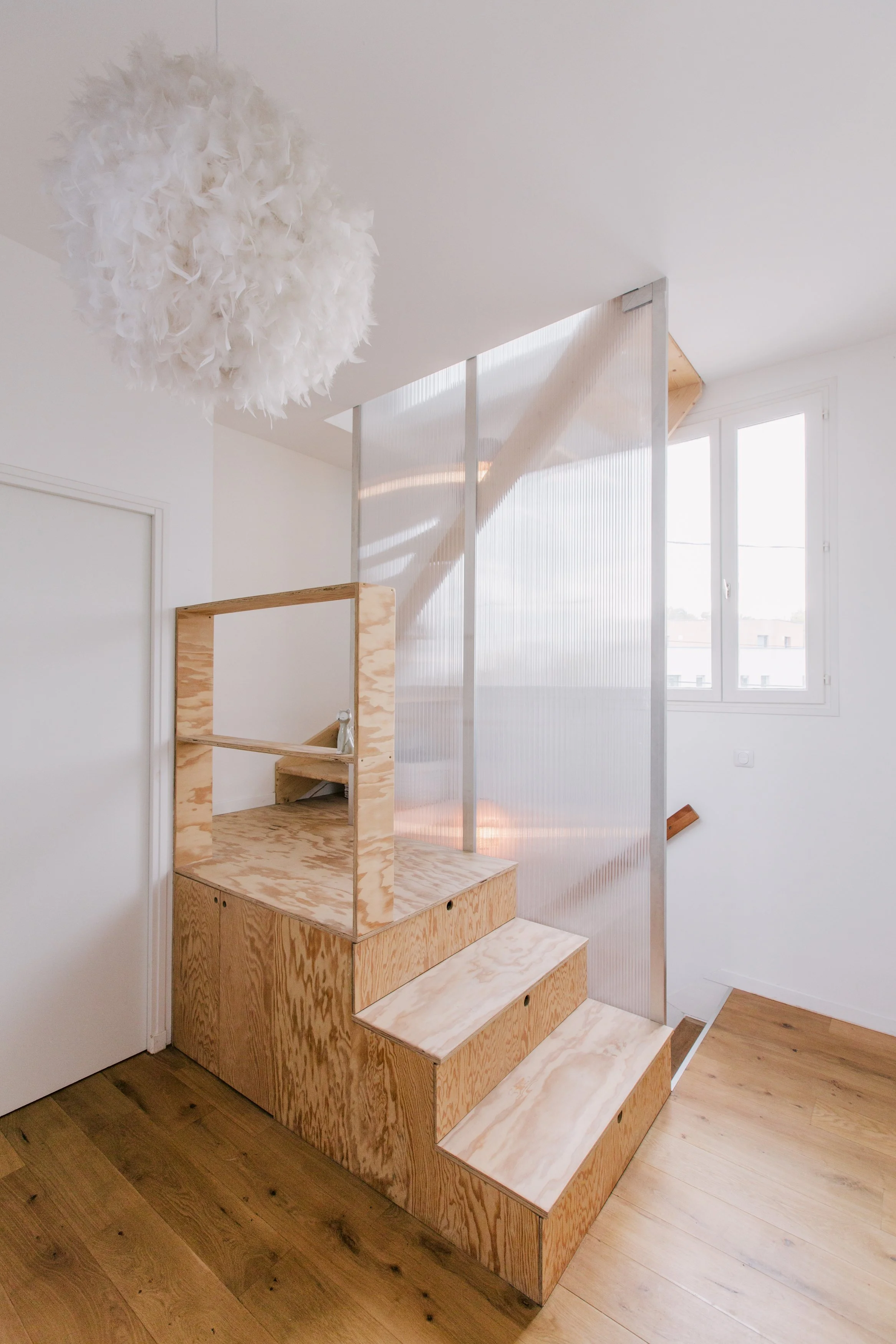 Aménagement intérieur avec un escalier en bois avec rangement intégré et une banquette, une cloison en plexiglas, fenêtres laissant entrer beaucoup de lumière naturelle, et un plafond avec un luminaire en forme de boule en plumes blanches.