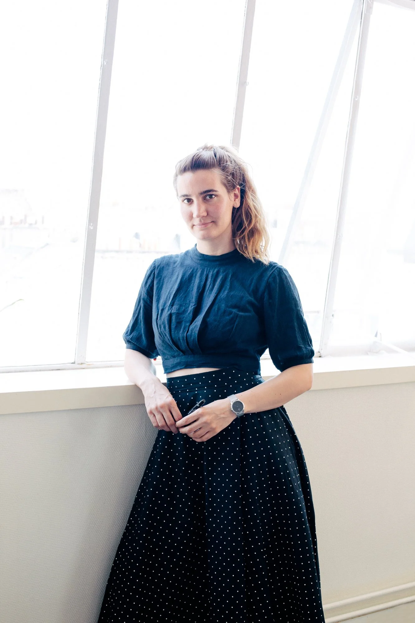 Femme debout près d'une grande fenêtre, portant une robe noire à pois blancs, regarde vers l'objectif, tenant un stylo, avec un fond lumineux