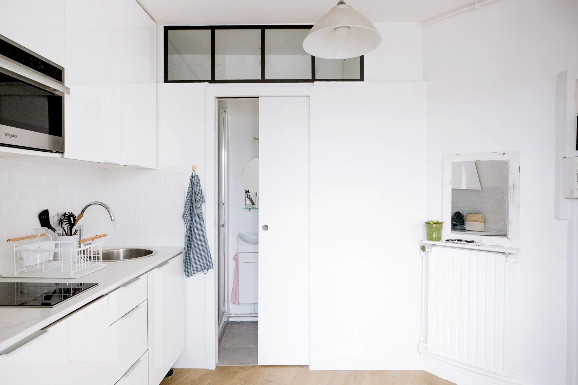 Cuisine blanche moderne avec plan de travail en marbre, évier, micro-ondes intégré, et porte menant à une petite pièce derrière. Espace organisé avec des ustensiles de cuisine et un rideau à carreaux bleu et blanc suspendu à la poignée de la porte.