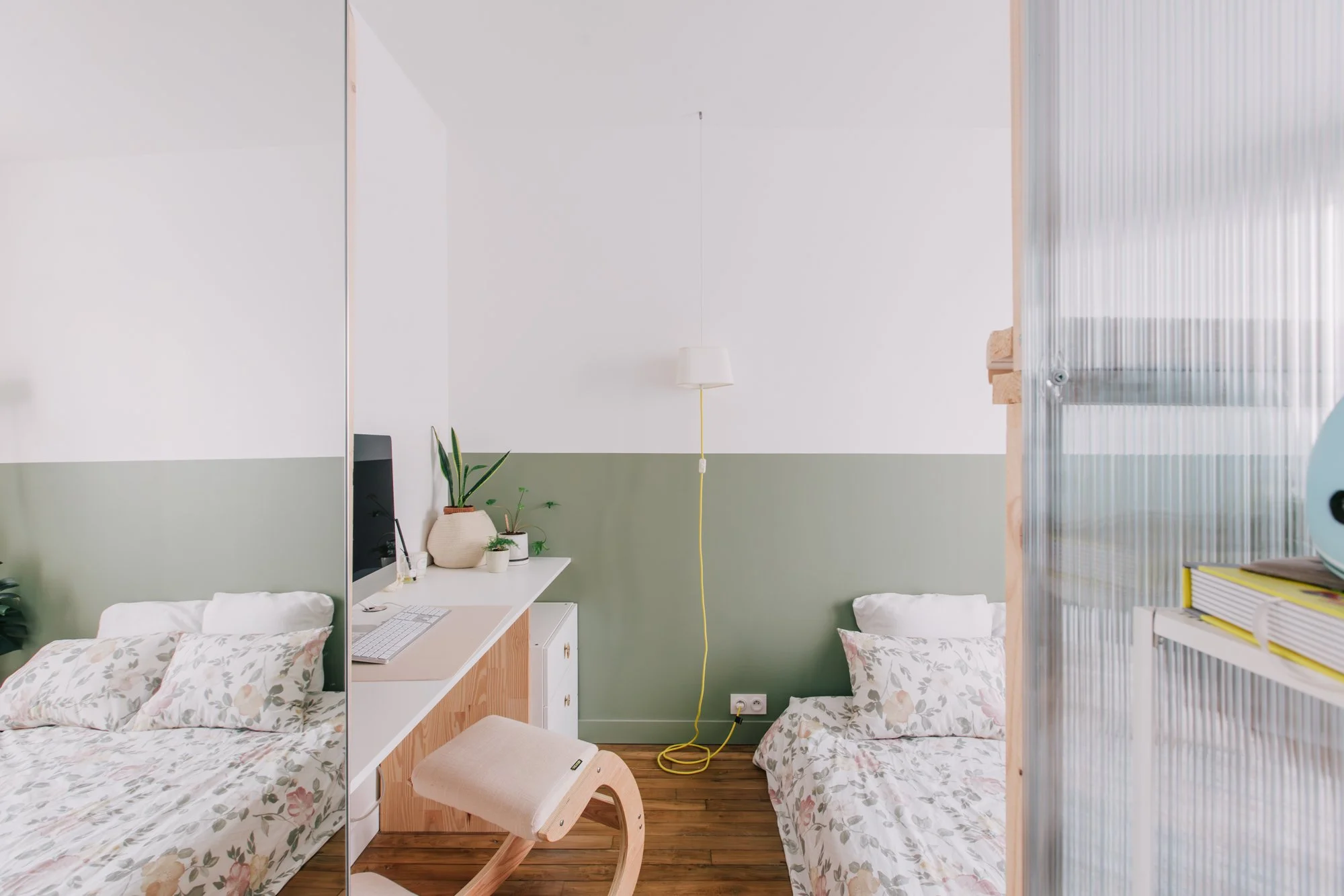 Chambre avec deux lits avec draps à fleurs, une étagère avec des plantes, un bureau blanc avec un ordinateur et une lampe suspendue, séparée par une cloison en plexiglas.