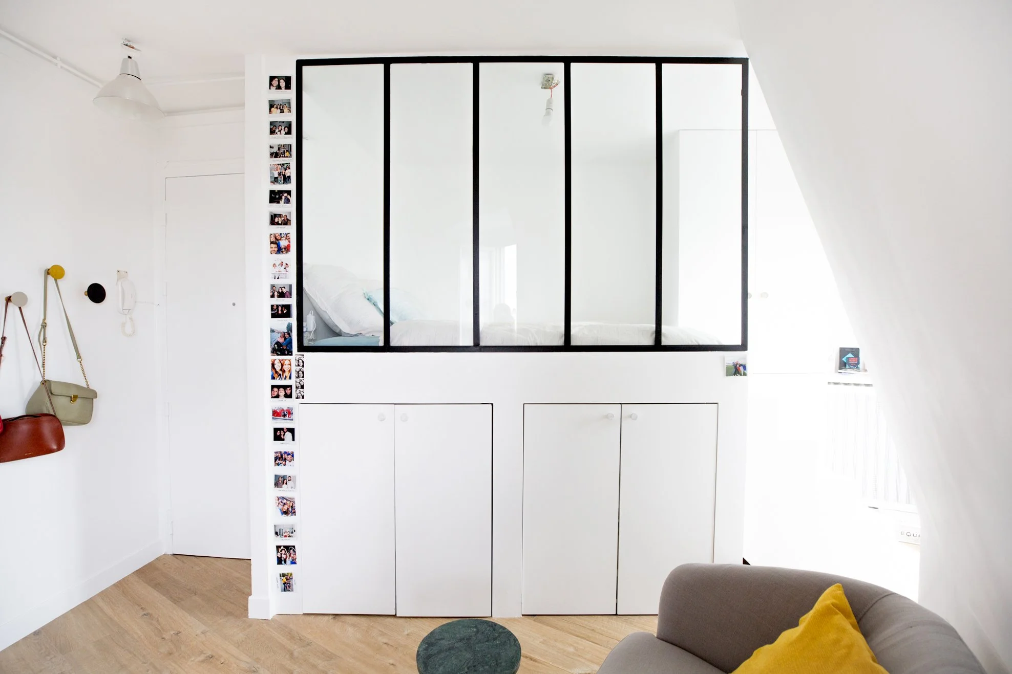 Intérieur d'un appartement avec un espace salon, mur blanc, avec une grande armoire blanche munie de portes, un coussin jaune sur un canapé, un tapis vert foncé et un coin avec des sacs suspendus et des photos en bande sur le mur.