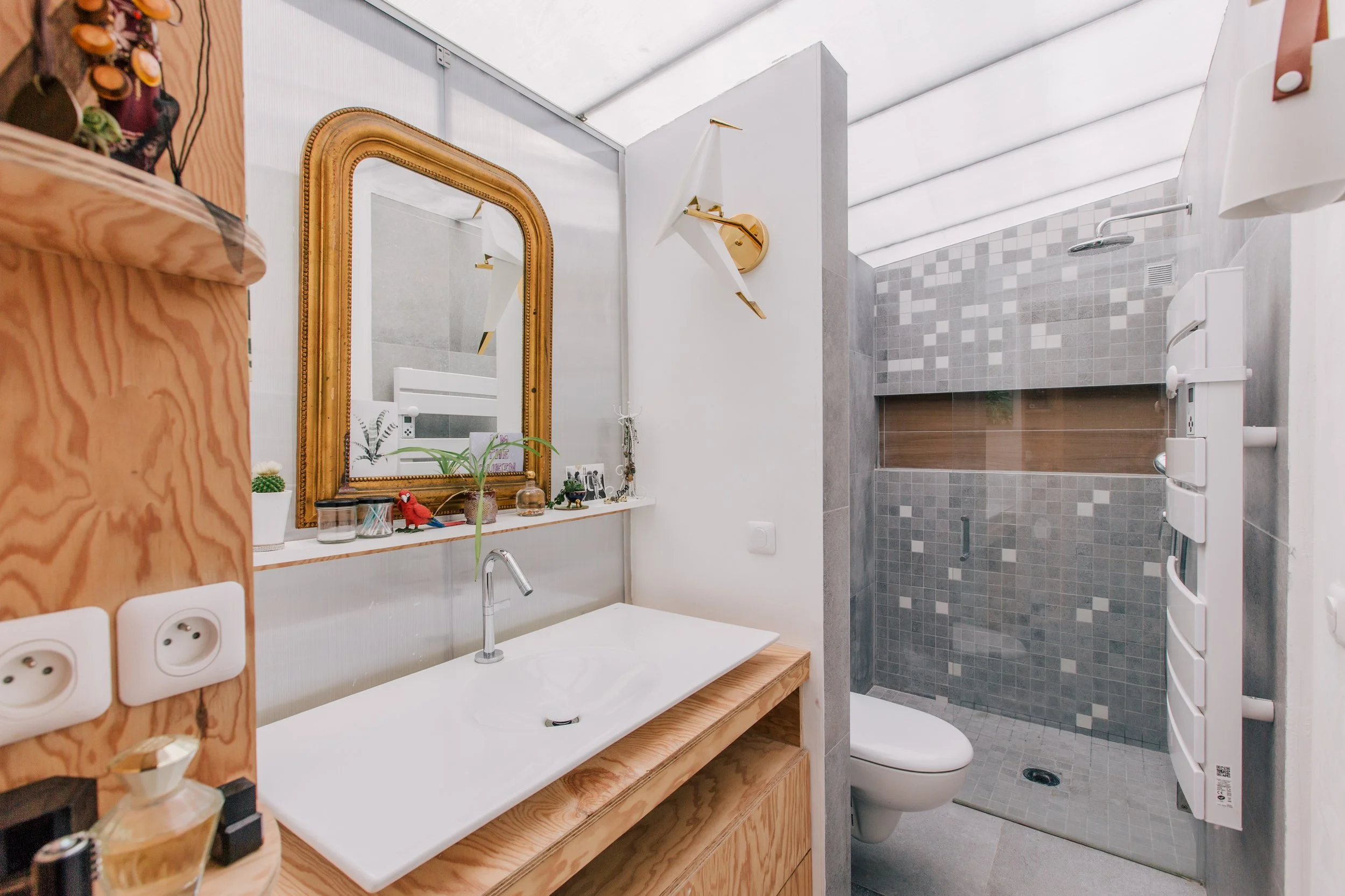 Une salle de bain moderne avec un miroir doré, une vasque blanche sur un meuble en bois, des décorations et des plantes, une séparation en verre menant à une douche murée en carreaux gris et un WC.