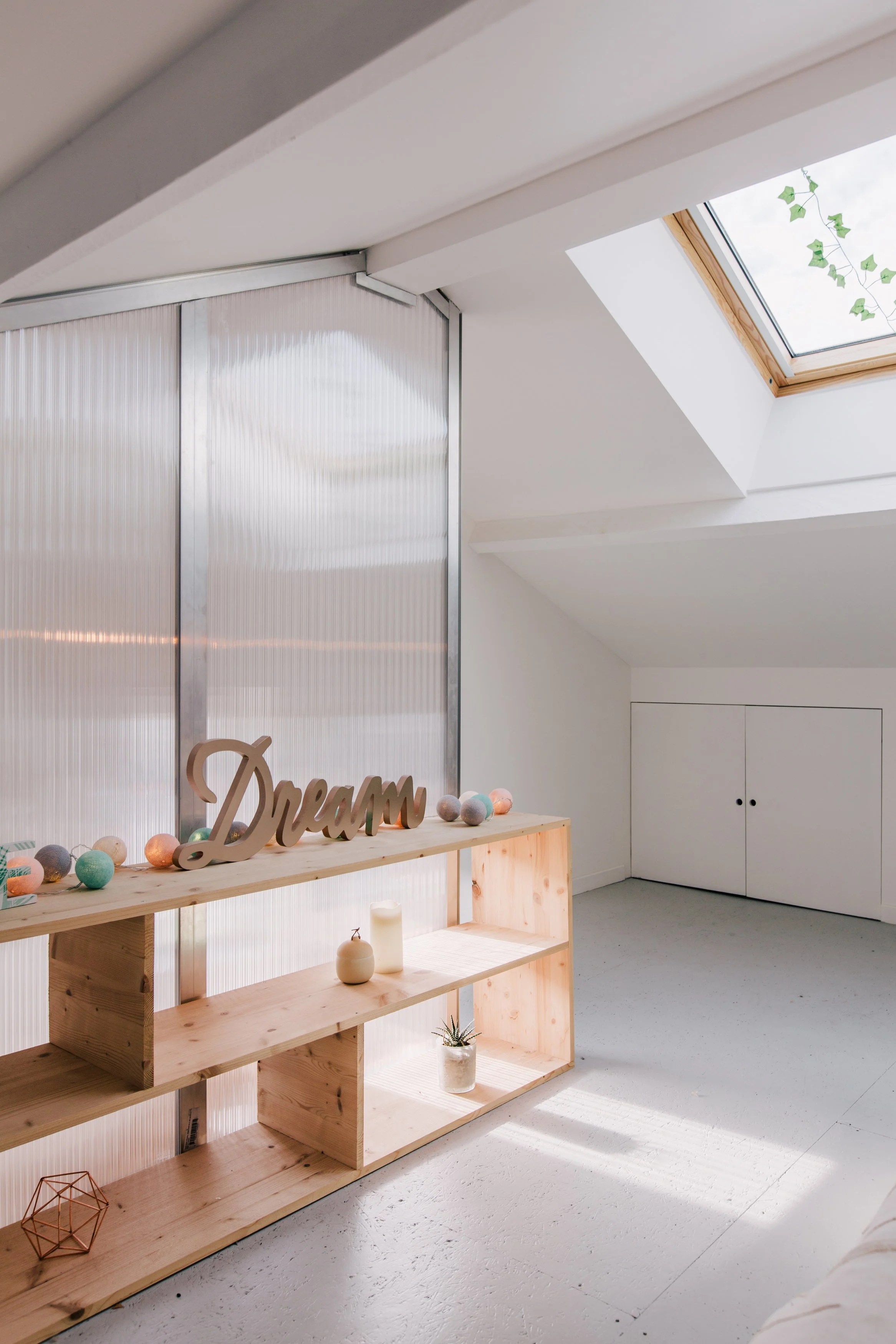 Une étagère en bois clair décorée de boules en laine colorées, d'une enseigne en bois formant le mot 'Dream', et de diverses petites décorations comme des bougies, un petit pot avec un cactus, et un cube en fil de cuivre.