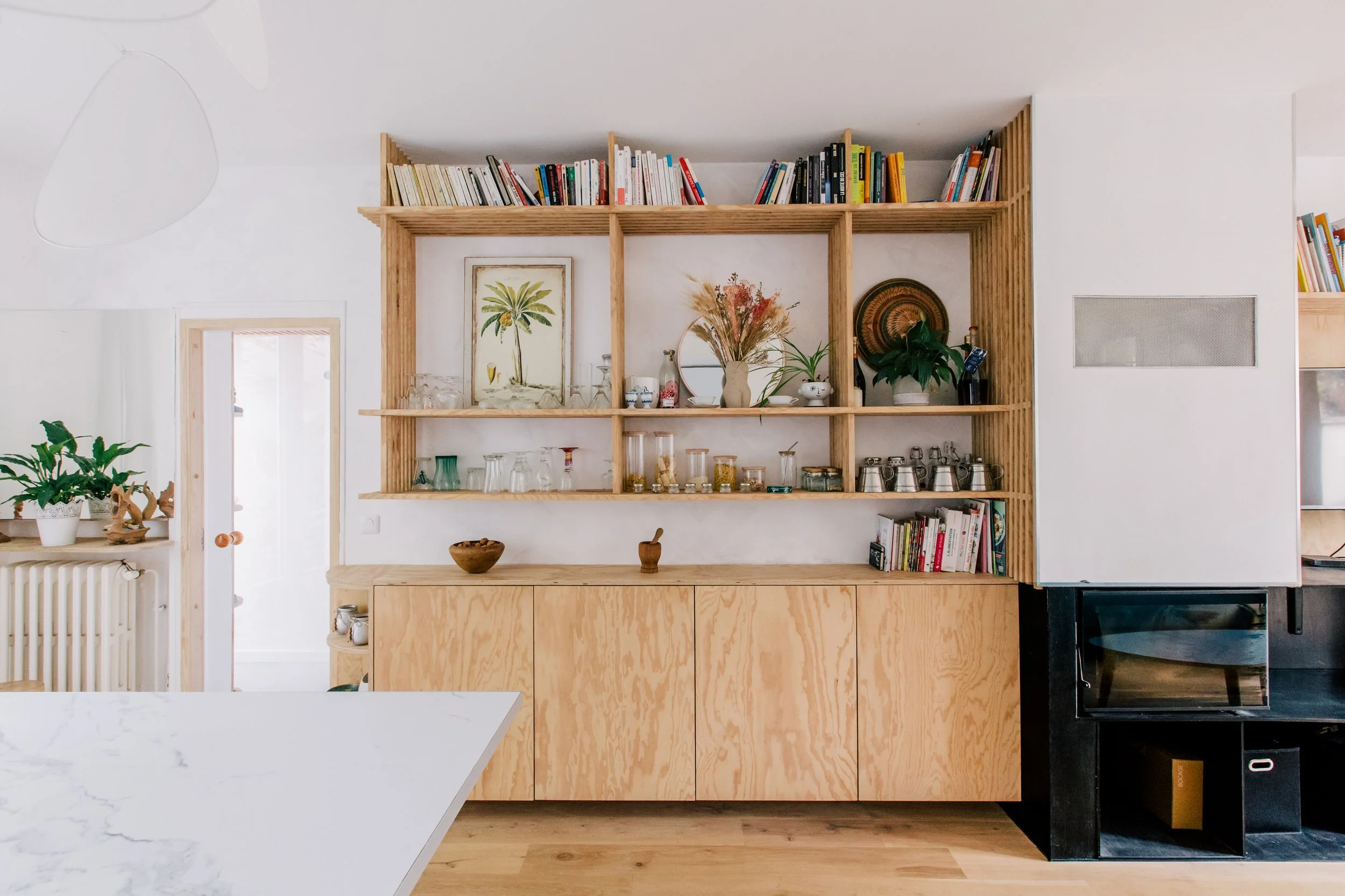 Une étagère en bois remplit le mur du salon, avec des livres, des plantes, des céramiques et des objets décoratifs. La pièce est lumineuse avec un style minimaliste.