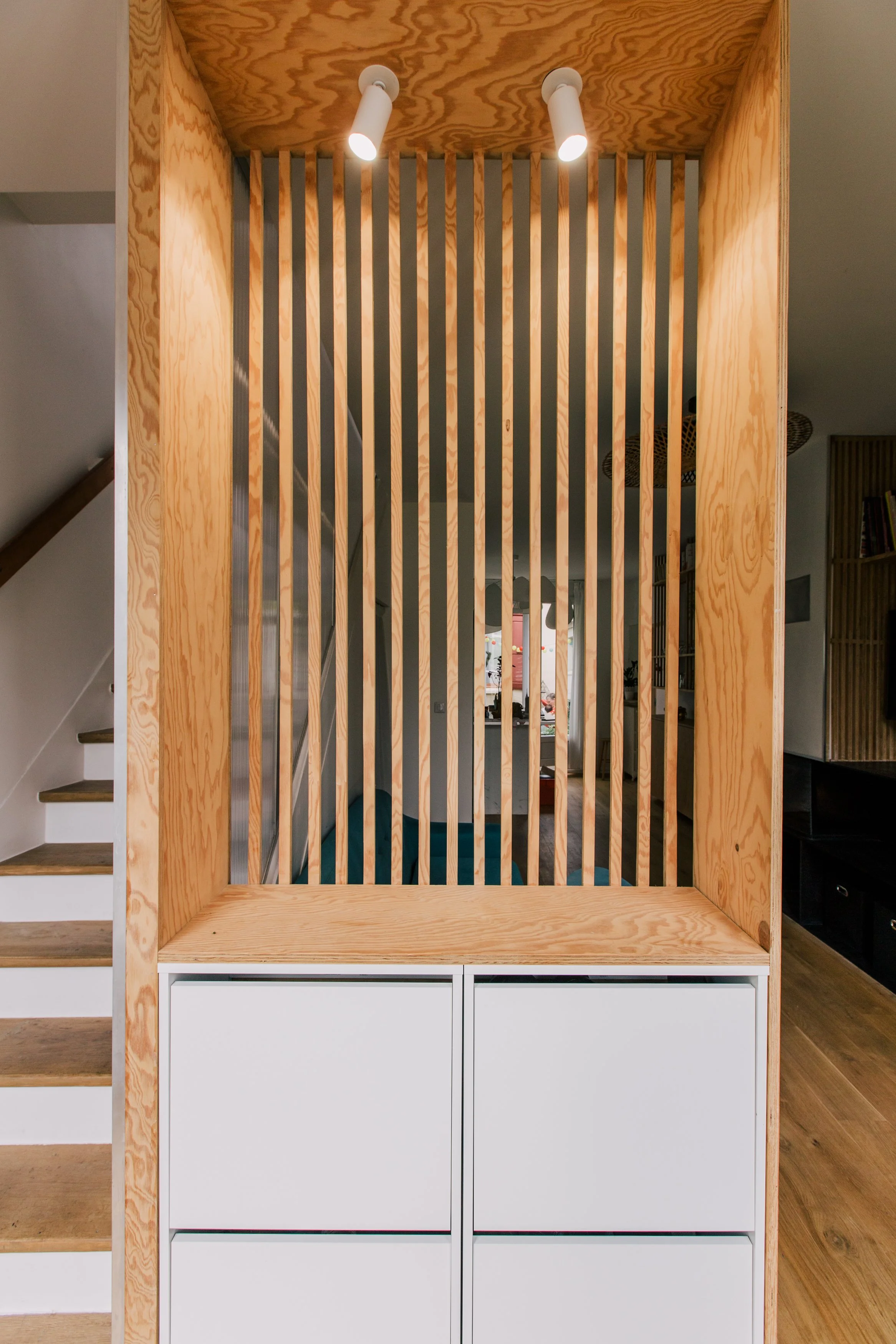 Un meuble en bois avec des barres verticales, éclairé par deux spots lumineux au plafond. En dessous, un meuble blanc avec des tiroirs.