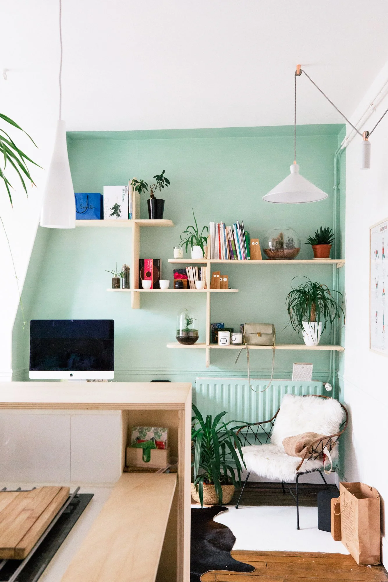Une salle de bureau avec un mur vert pâle, des étagères en bois blanc contenant des livres, des plantes, des décorations, une imprimante, et une chaise avec un fauteuil en peluche blanc. Il y a aussi des objets comme des paniers en osier, des sacs en