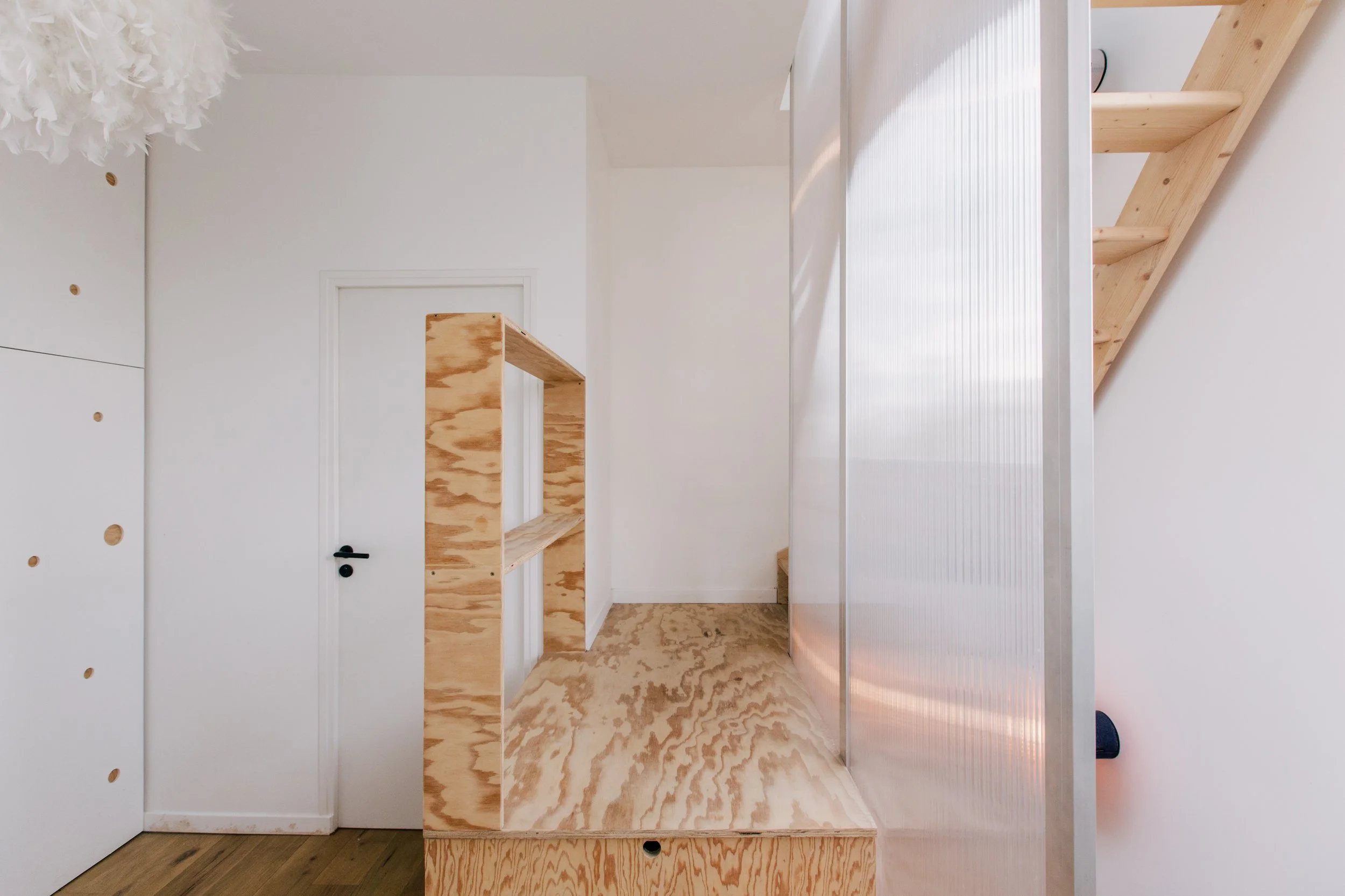 Salle blanche avec des meubles en bois non finis, un mur blanc, un plafond avec lampes, et un sol en bois.