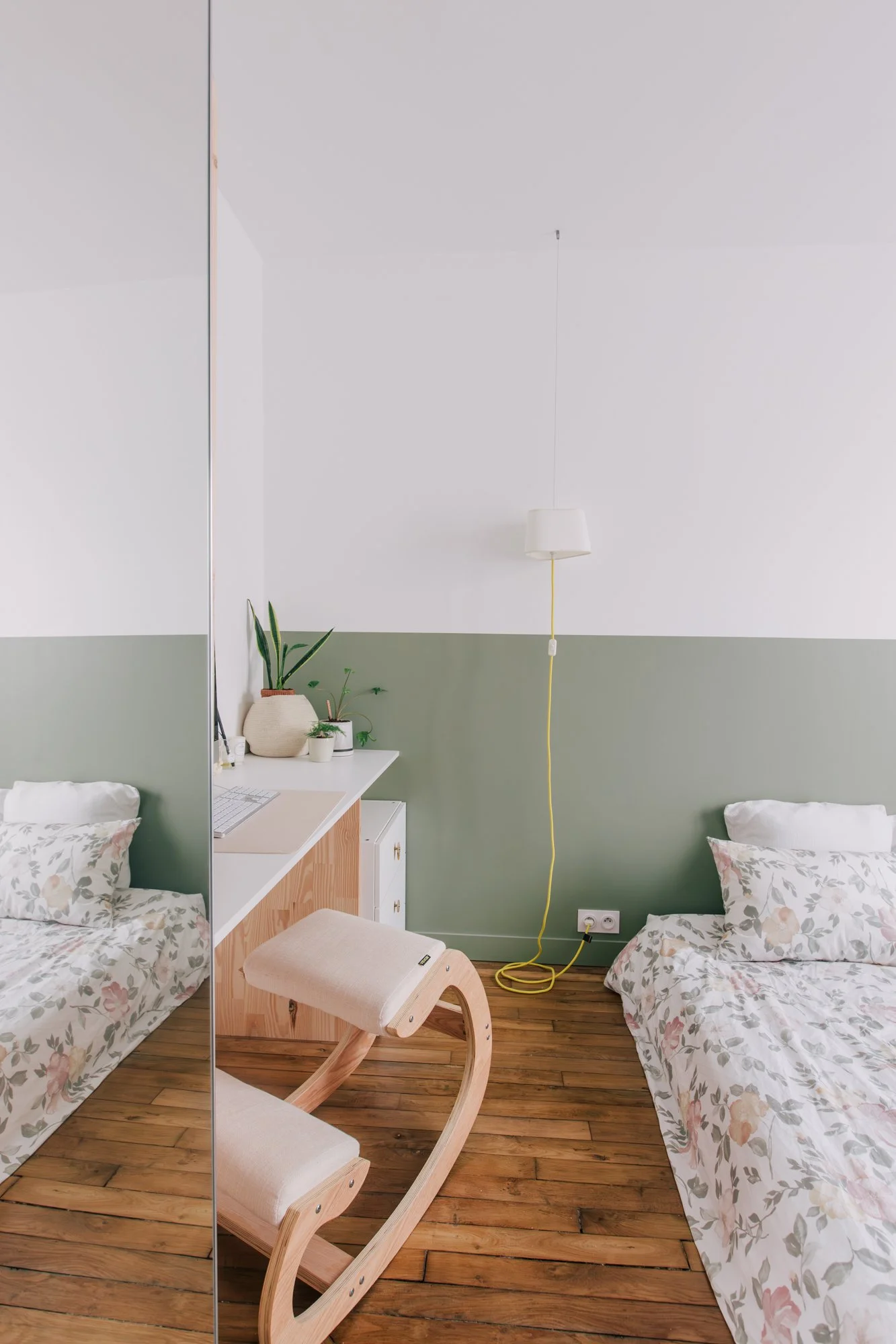 Chambre minimaliste avec deux lits, un bureau avec des plantes, un miroir, une lampe suspendue, et un fauteuil en bois, avec un sol en bois.