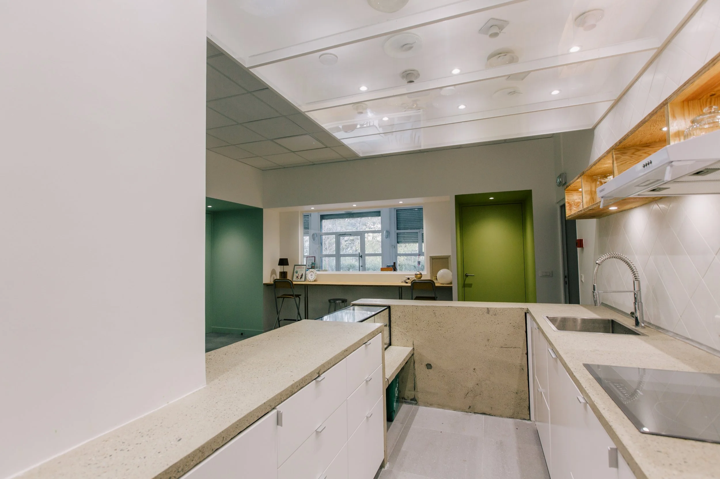 Cuisine moderne avec comptoirs en béton, armoires blanches et étagères en bois, fenêtres. Niches colorées.