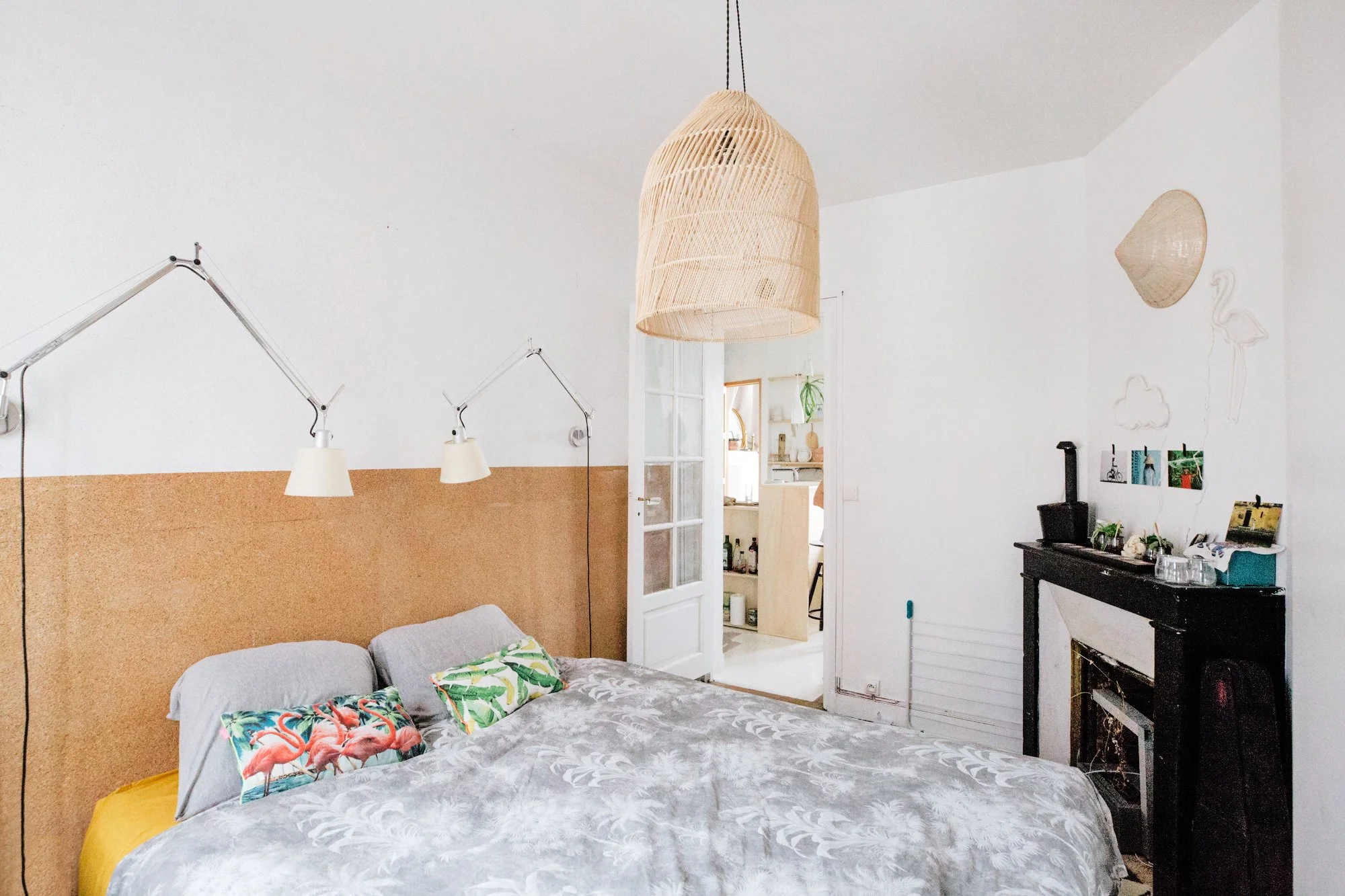 Chambre chaleureuse avec un lit double, oreillers à motifs de flamants roses, lampes de chevet. Déco minimaliste avec mur blanc et un mur en liège, et une suspension en rotin au plafond.