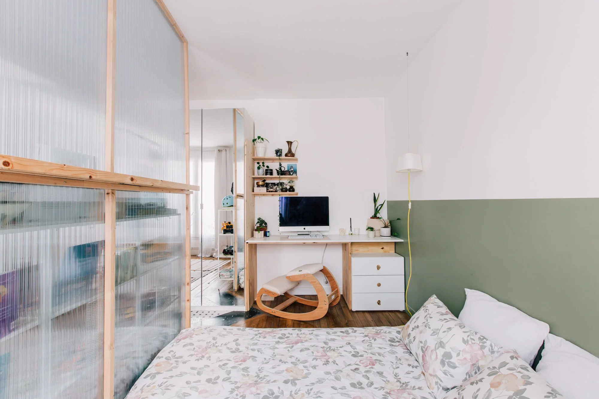 Chambre avec lit à fleurs, mobilier en bois, bureau avec ordinateur, étagères avec plantes et objets décoratifs, paroi en plastique translucide.