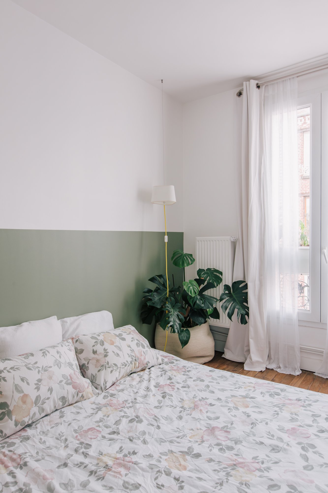Chambre avec lit double, oreillers à fleurs, mur blanc avec partie verte, grande plante verte, lampe suspendue, rideaux blancs, fenêtre ouverte, parquet en bois