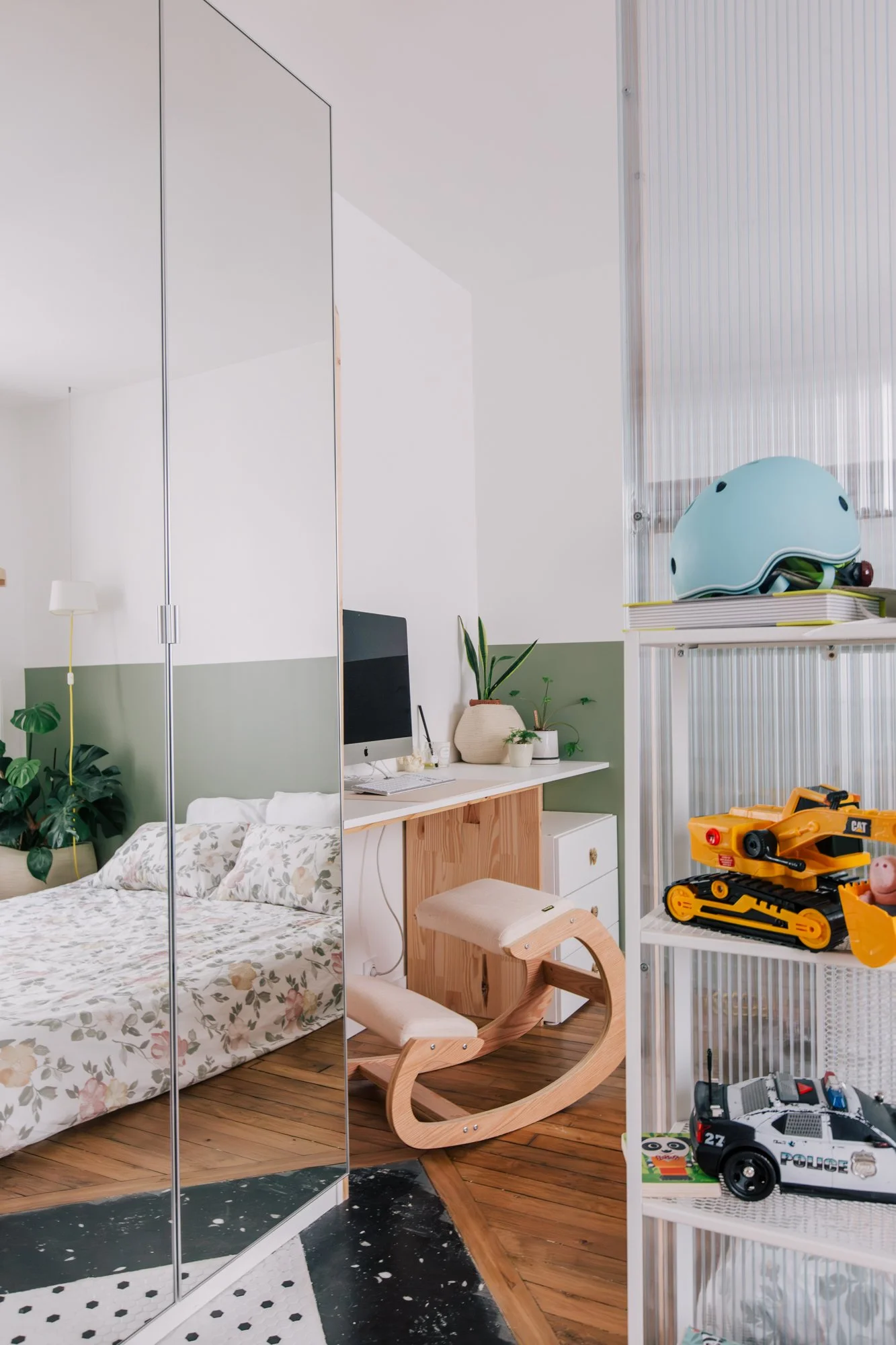 Intérieur d'une chambre moderne avec un mur en miroir, un bureau avec un ordinateur, des plantes, une chaise en bois, étagère avec jouets pour enfants, y compris une voiture de police, un casque de vélo, et un tapis noir et blanc.