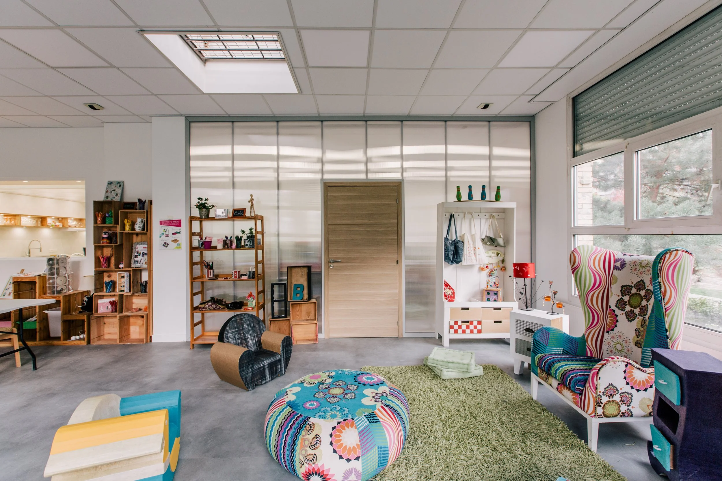 Intérieur d'une salle colorée avec des meubles en bois, une chaise à motifs, des étagères avec des objets décoratifs et des jouets, grande fenêtre laissant entrer la lumière naturelle.