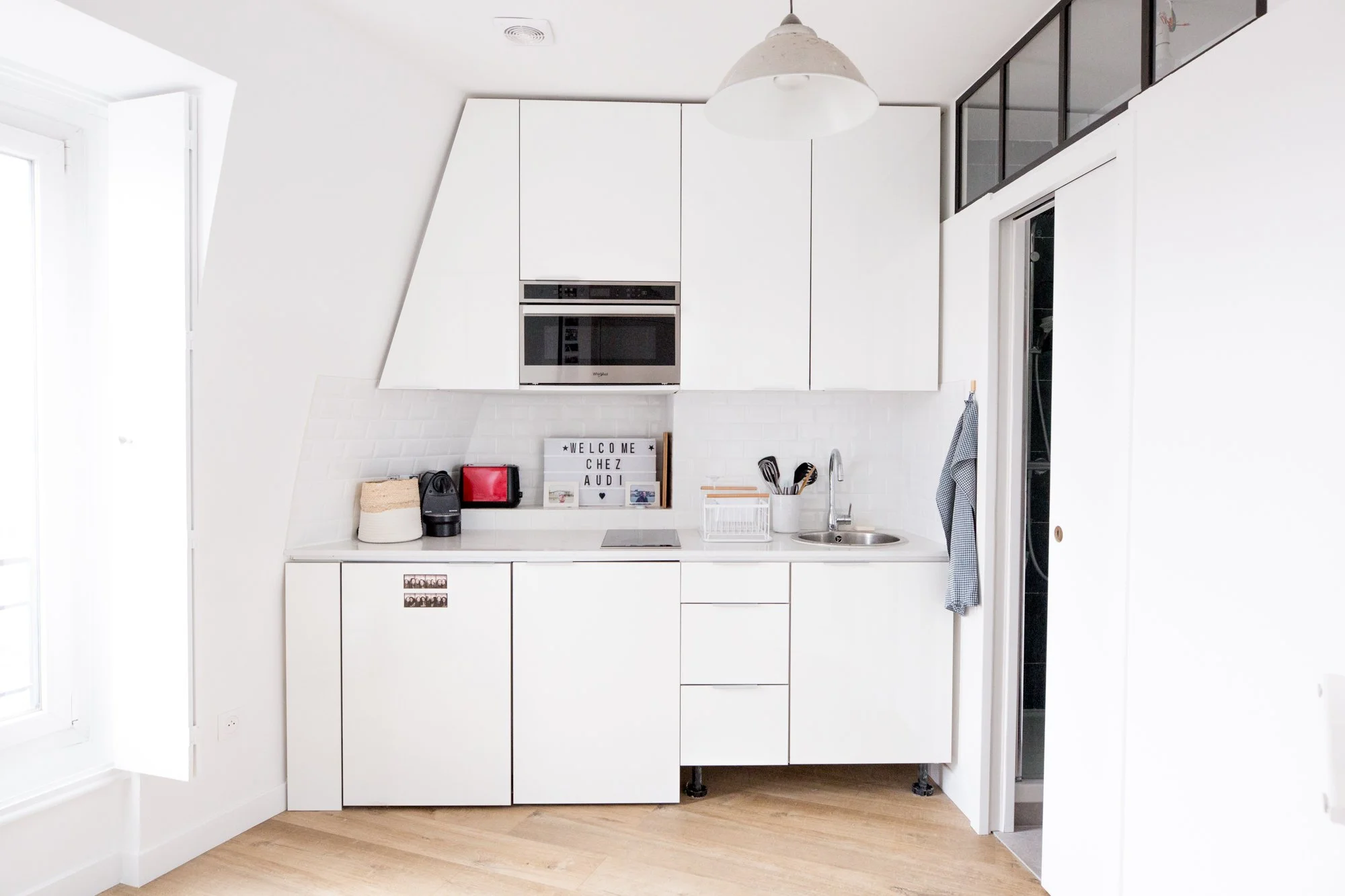 Cuisine moderne blanche avec évier, micro-ondes, étagère décorative, et une grande fenêtre à gauche.