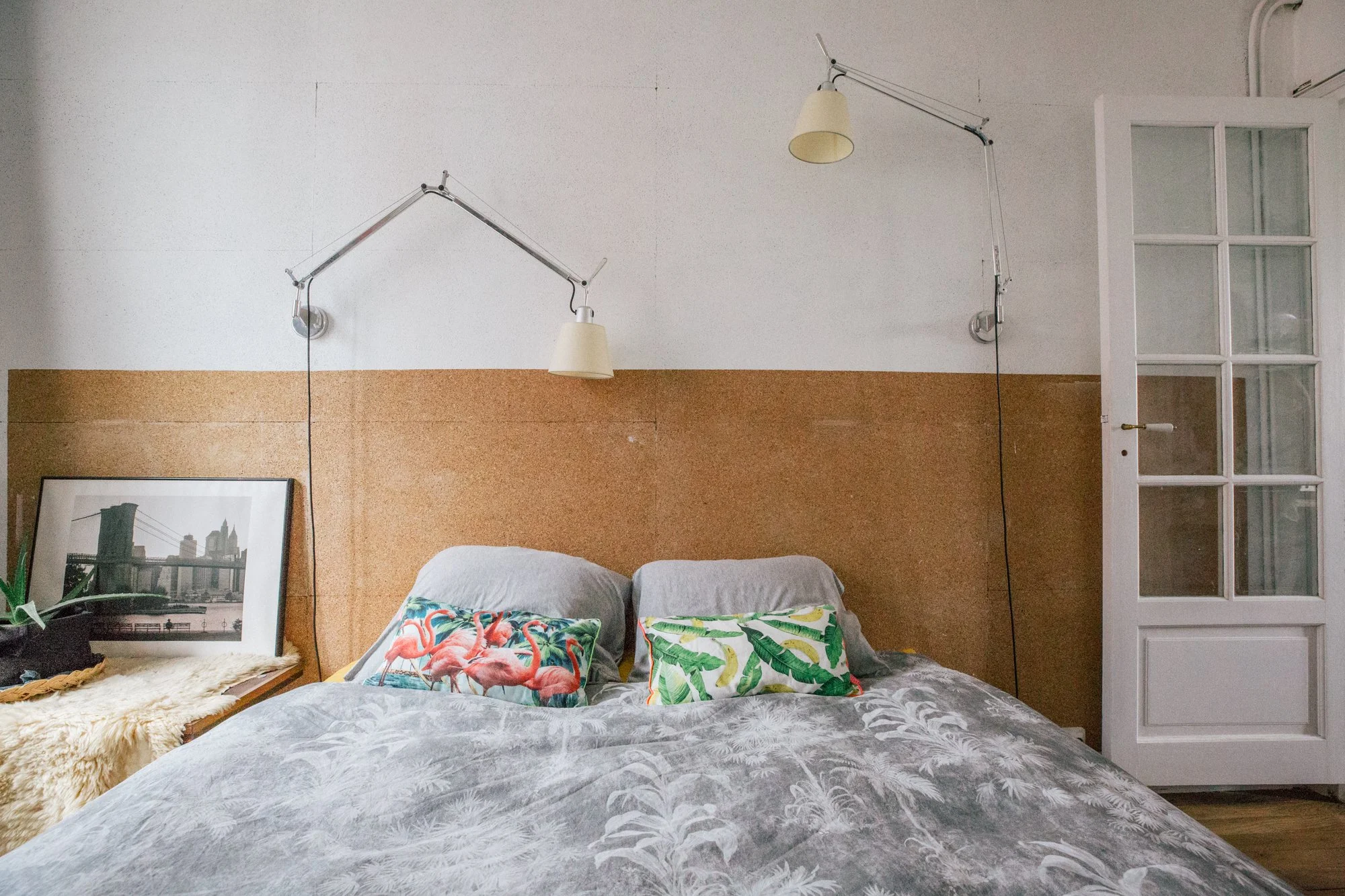 Un lit avec deux oreillers décorés de motifs de flamingos et de feuilles tropicales, dans une chambre au mur en bois clair, avec une affiche encadrée à gauche et des lampes murales au-dessus.
