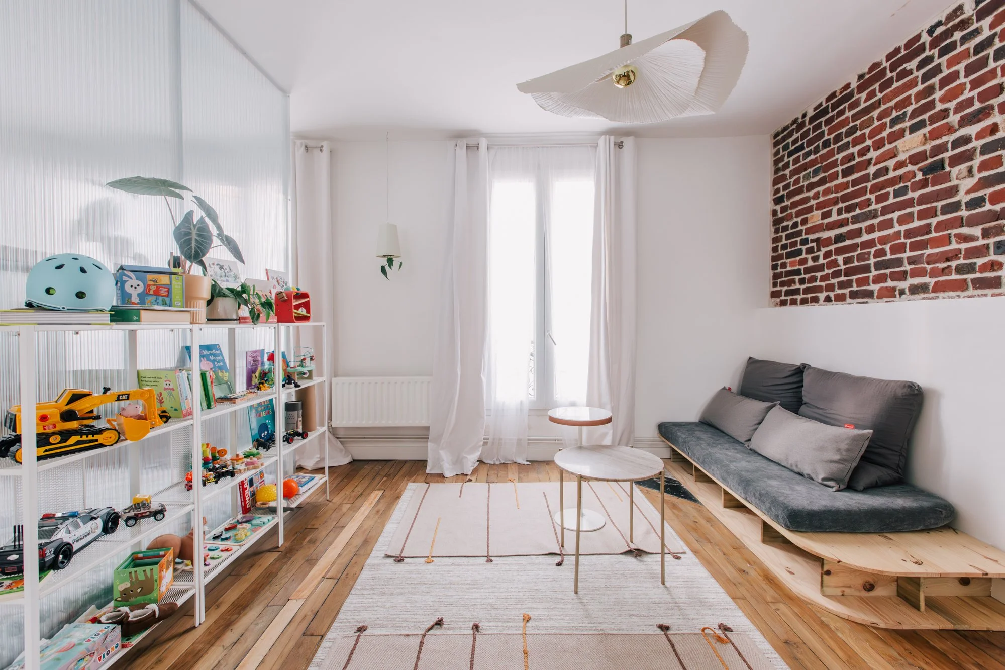 Salon avec étagère remplie de jouets pour enfants, canapé gris avec coussins, table basse ronde, tapis beige et décor moderne avec mur en briques rouges et grandes fenêtres avec rideaux blancs.