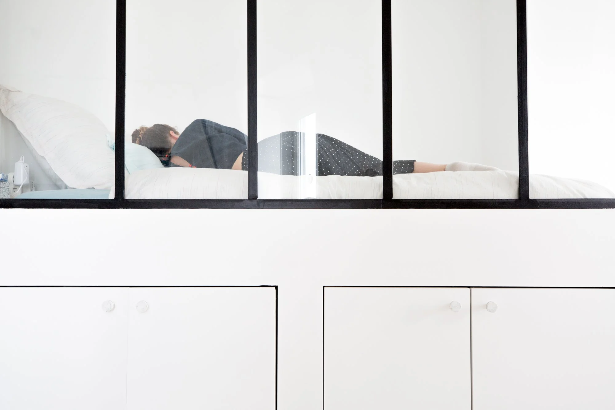 Une personne repose dans un lit dans une pièce blanche, vue de la fenêtre ou d'une paroi en verre avec cadres noirs.