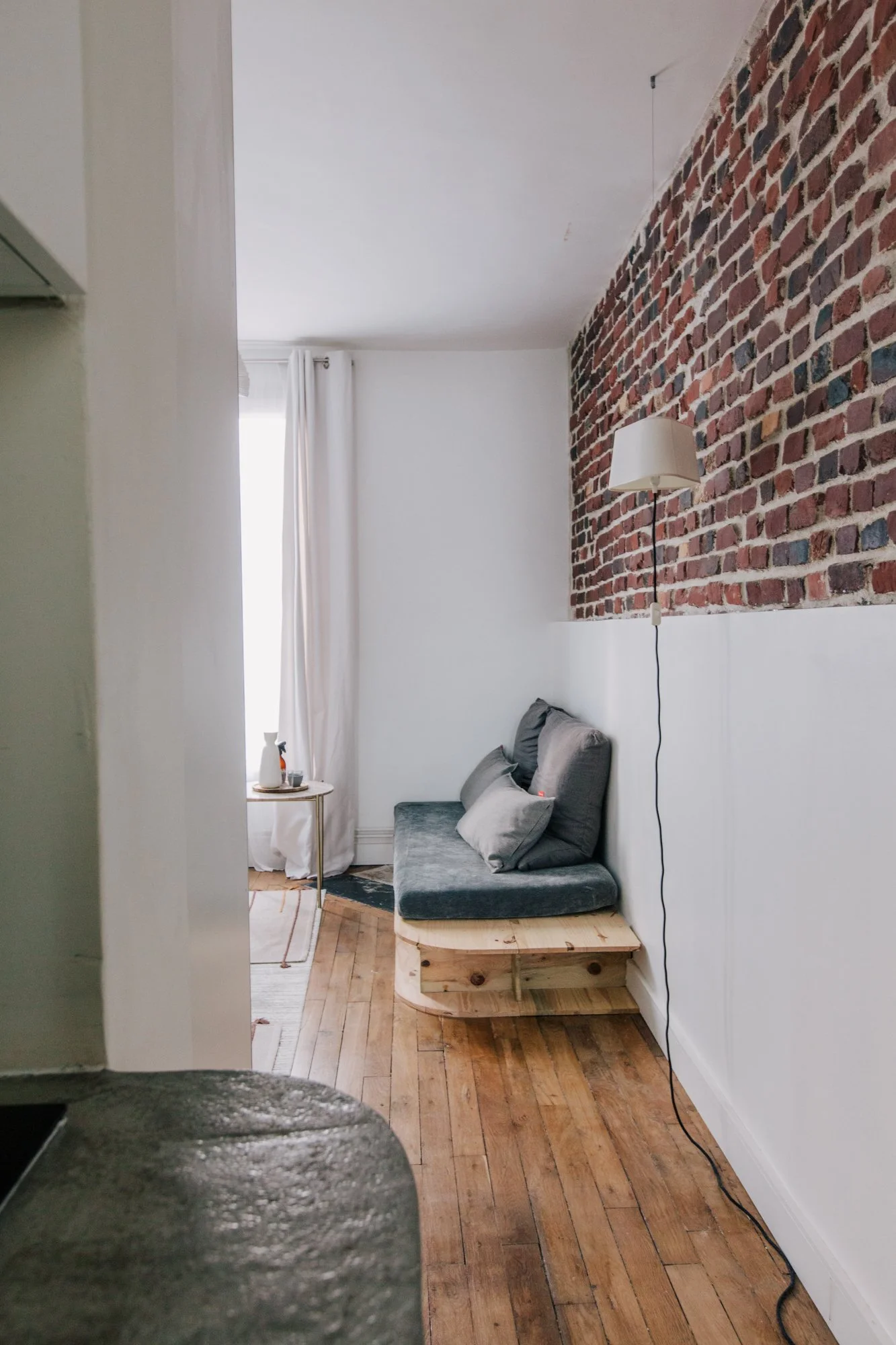 Intérieur d'un salon avec un mur en briques rouges, un canapé gris sur une plateforme en bois, une lampe murale, une fenêtre avec rideaux blancs, et un sol en parquet en bois.