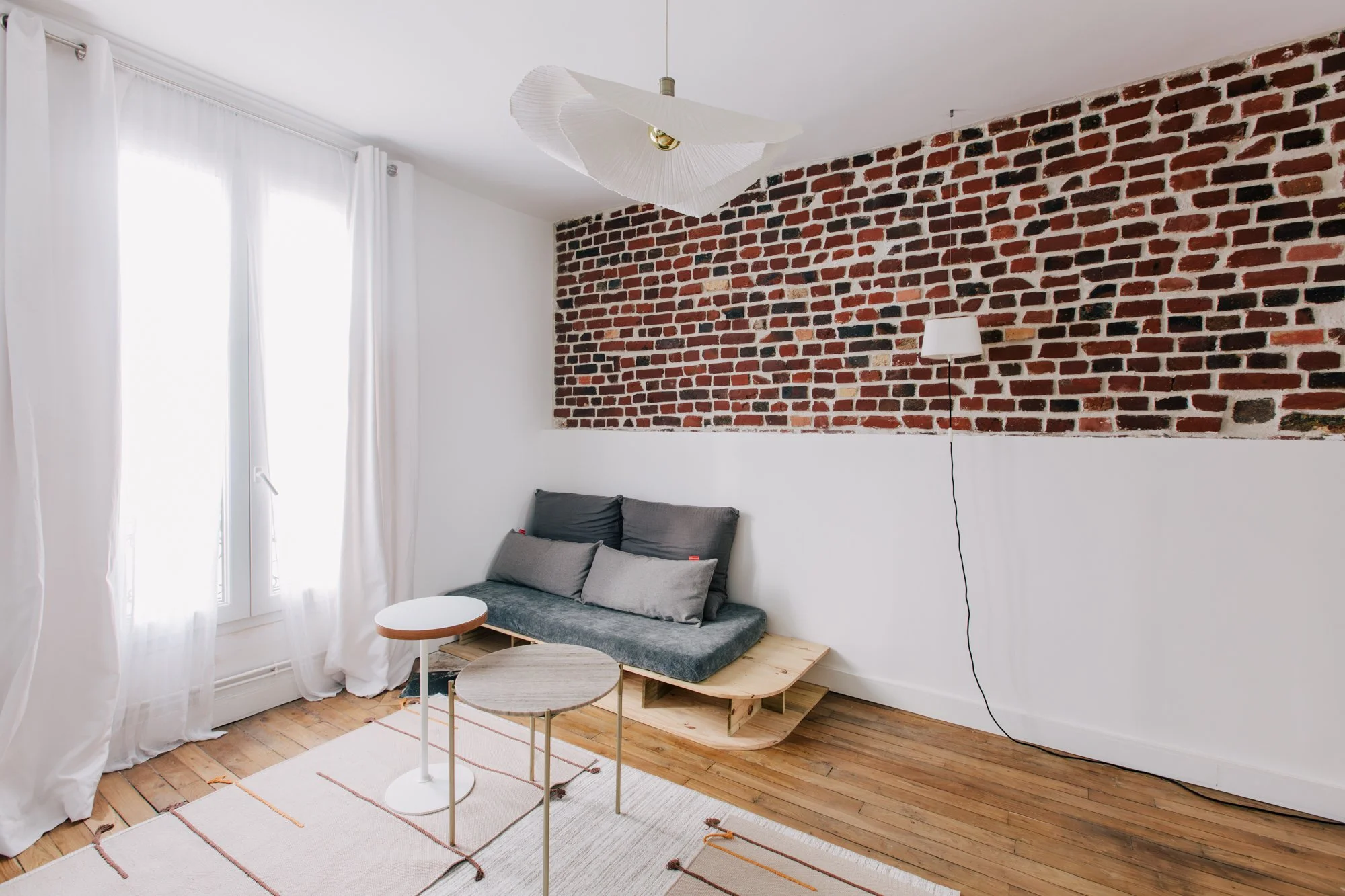 Salon avec une brique rouge sur le mur, un canapé en dessous de la brique, deux tables rondes, une lampe suspendue, une lampe mural, des rideaux blancs, un sol en bois, un tapis blanc avec des lignes orange, et un canapé décoré de coussins gris.