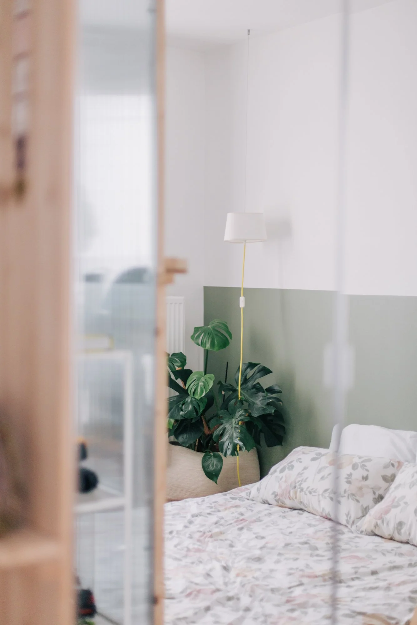 Intérieur d'une chambre avec un lit, une plante verte, un lampadaire blanc, et un mur blanc avec une zone grise, vue partielle à travers une porte ou un mur en bois.