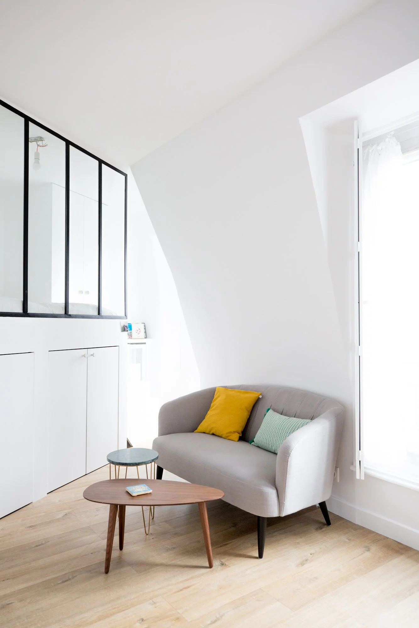 Salon intérieur moderne avec un canapé gris, un coussin jaune, et un coussin vert, deux tables basses en bois, un mur blanc avec une porte-fenêtre laissant entrer la lumière, une étagère blanche au mur, un plafond en pente, parquet en bois clair.