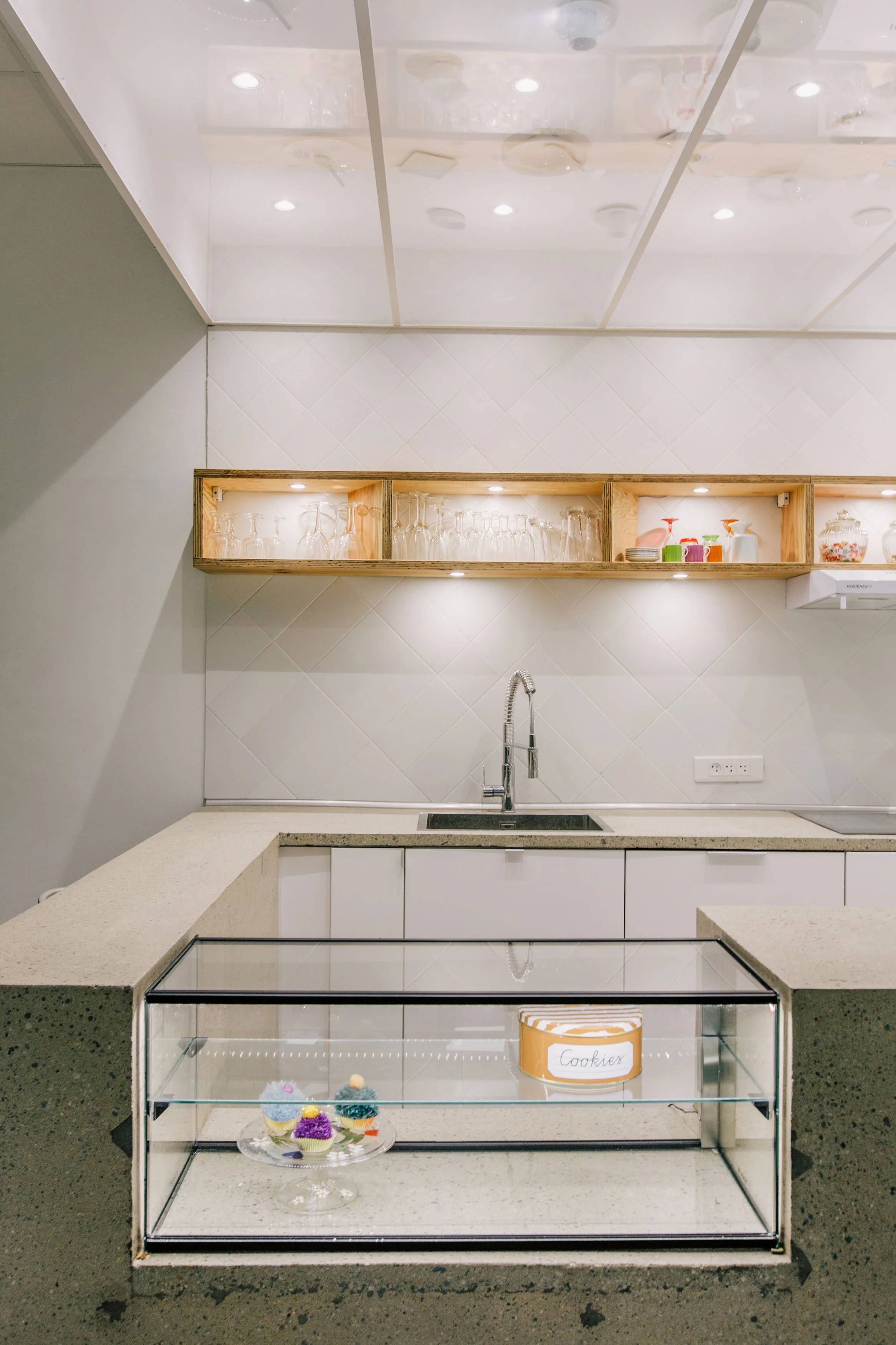 Une vitrine à cookies dans une cuisine moderne. La vitrine en verre affiche des cupcakes colorés, et un pot étiqueté 'Cookies' est placé à l'intérieur de la vitrine. Une étagère en bois au-dessus du lavabo contient des verres et des mugs.