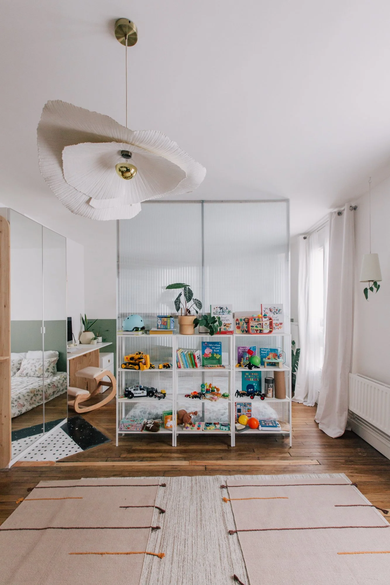 Une pièce d'enfant bien rangée avec des jouets, une étagère avec des livres et jouets, un lit avec un lit de bébé, des tapis sur le sol, une grande fenêtre avec rideaux, et un plafonnier blanc décoratif.