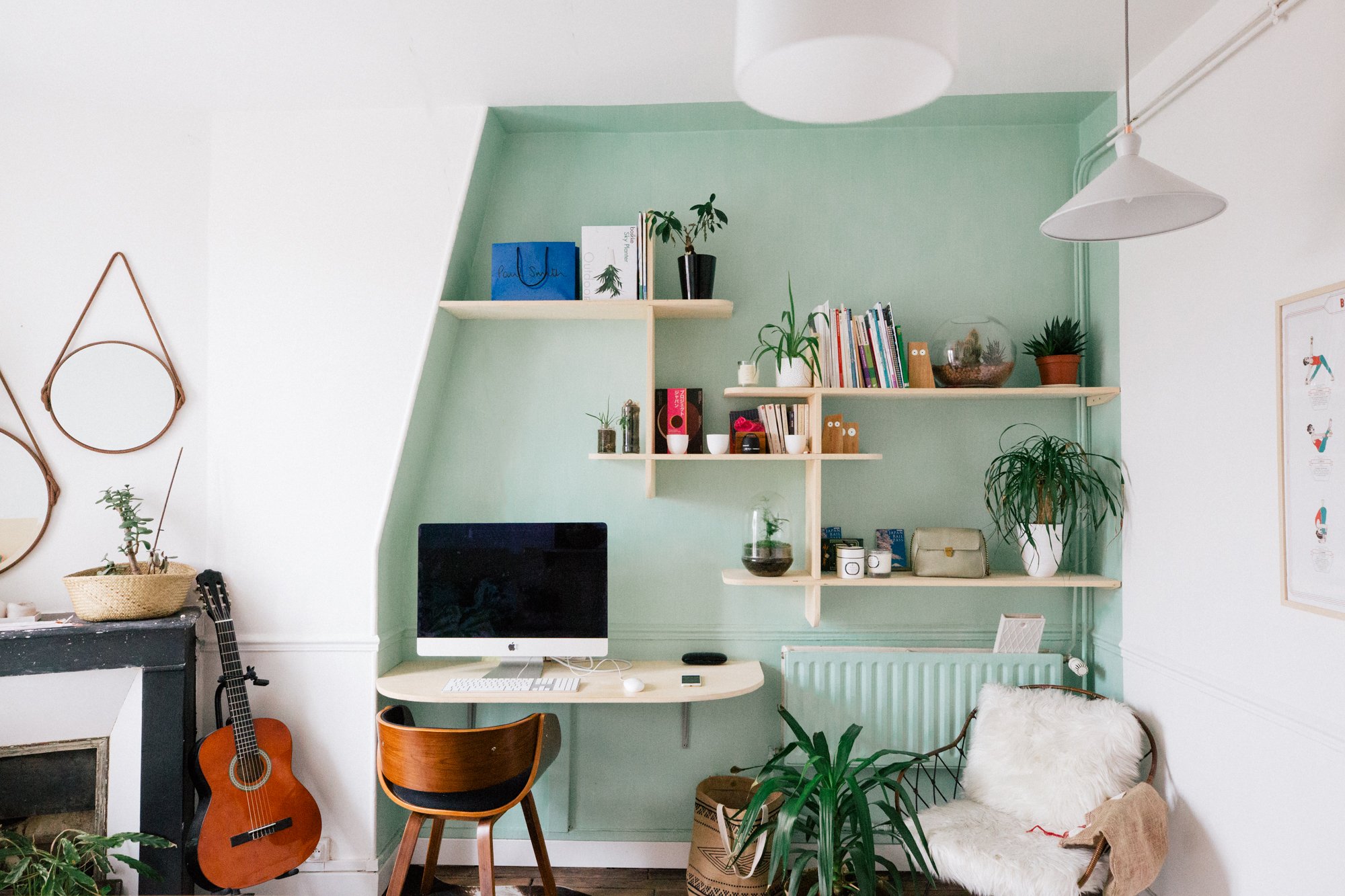 Un bureau avec un ordinateur iMac, une chaise en bois, et une étagère avec des livres, des plantes et des décorations. Une guitare est posée contre un meuble noir, et un fauteuil avec une couverture en fourrure blanche.