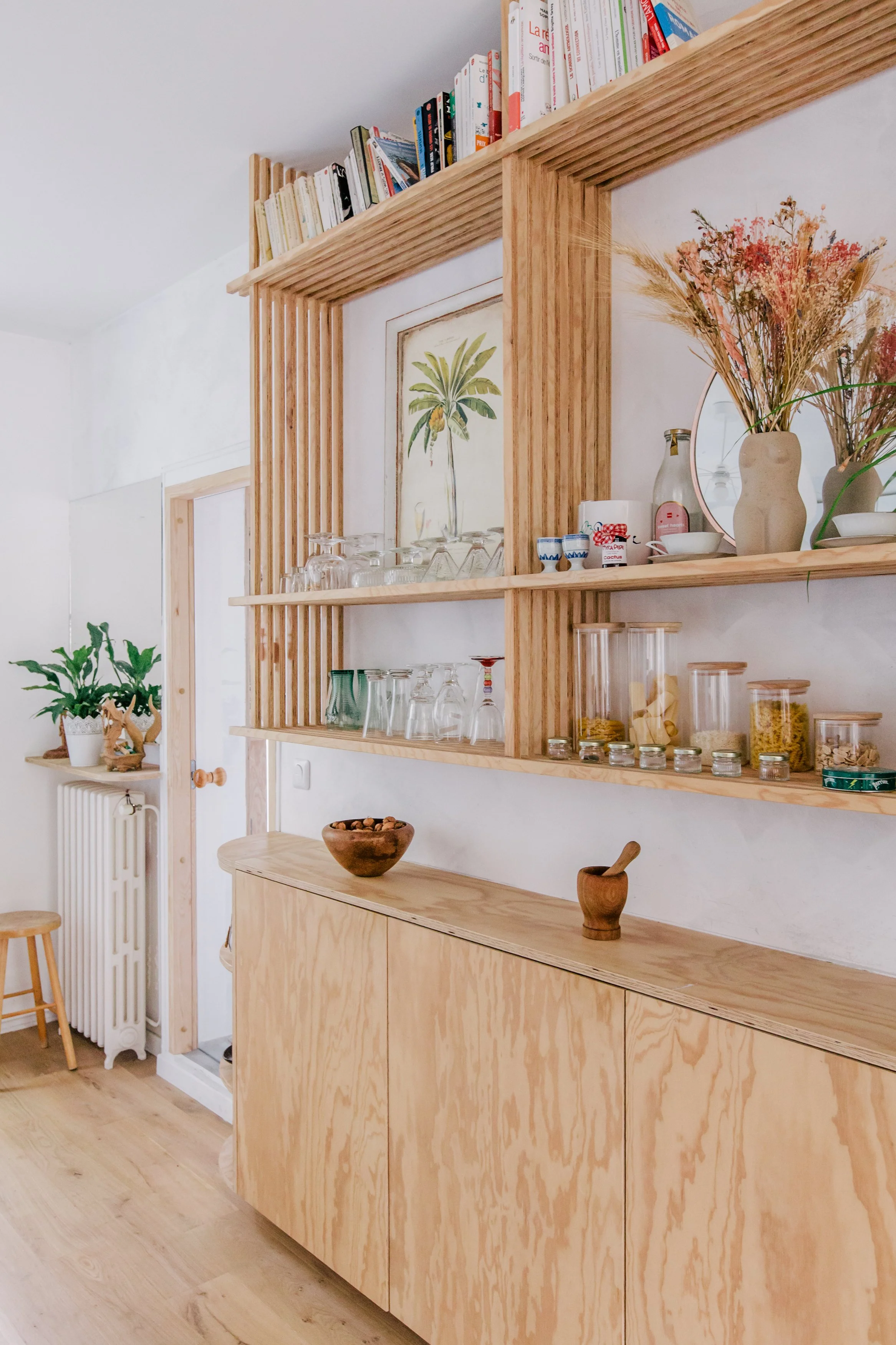 Une étagère en bois clair avec des livres posés en haut, des verres et des pots en verre avec des aliments en dessous, un miroir rond avec des fleurs séchées en vase, une peinture de palmier au mur, une plante verte en pot à gauche, un meuble en bois