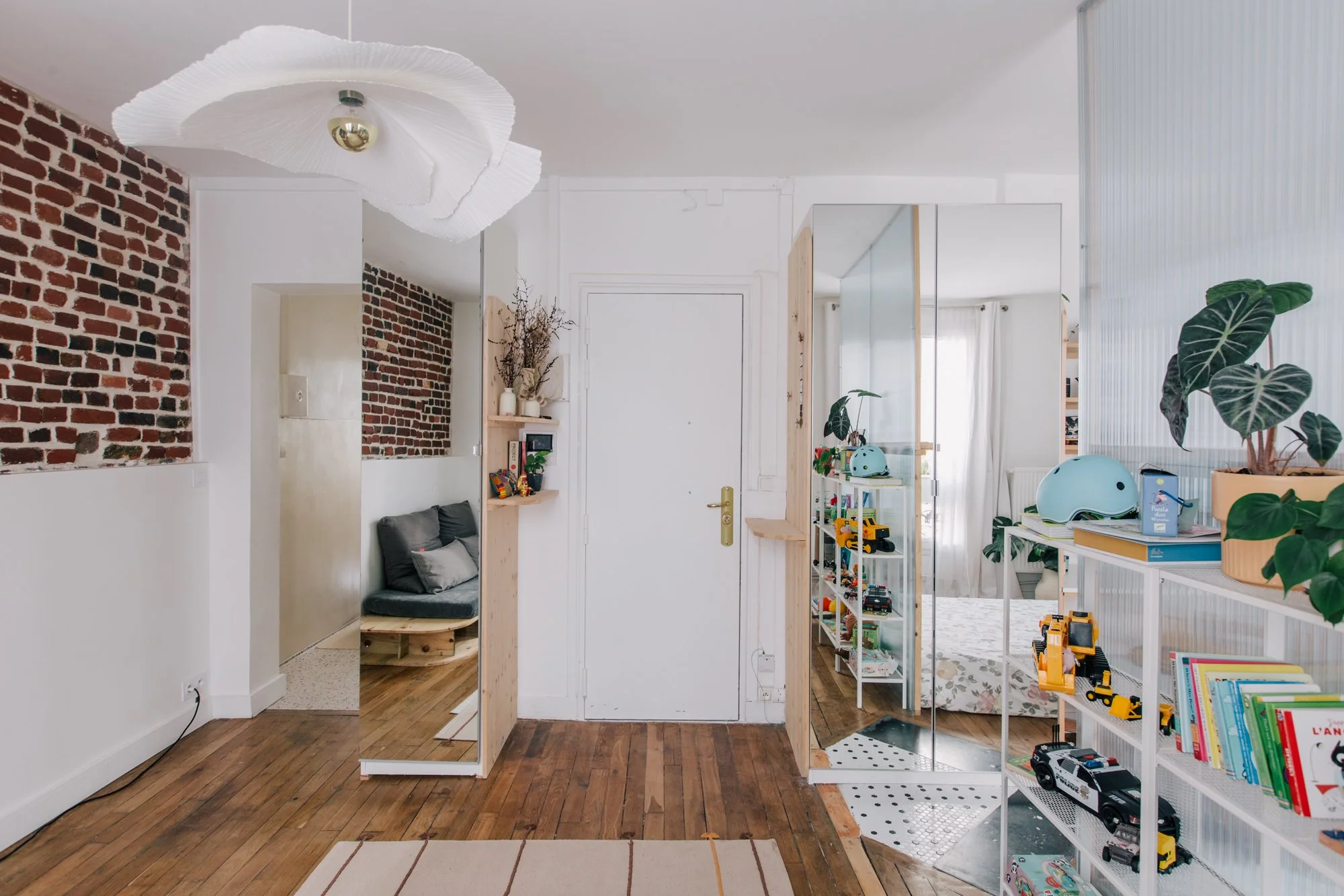 Intérieur d'un salon ou salle de jeux avec parquet en bois, murs blancs, décor moderne, et jouets pour enfants, plantes vertes, miroir, et meubles en bois clair.