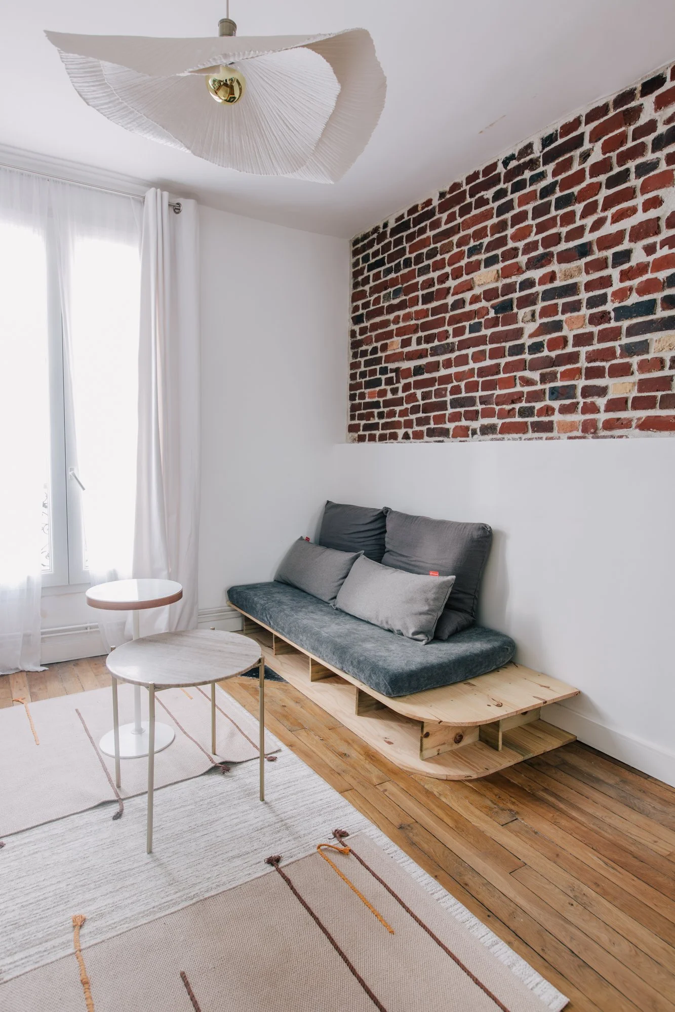 Salon avec un canapé en bois et tissu gris, deux tables basses, un tapis beige à rayures, une fenêtre avec voilages blancs, un mur en briques apparentes, un plafond blanc avec un luminaire