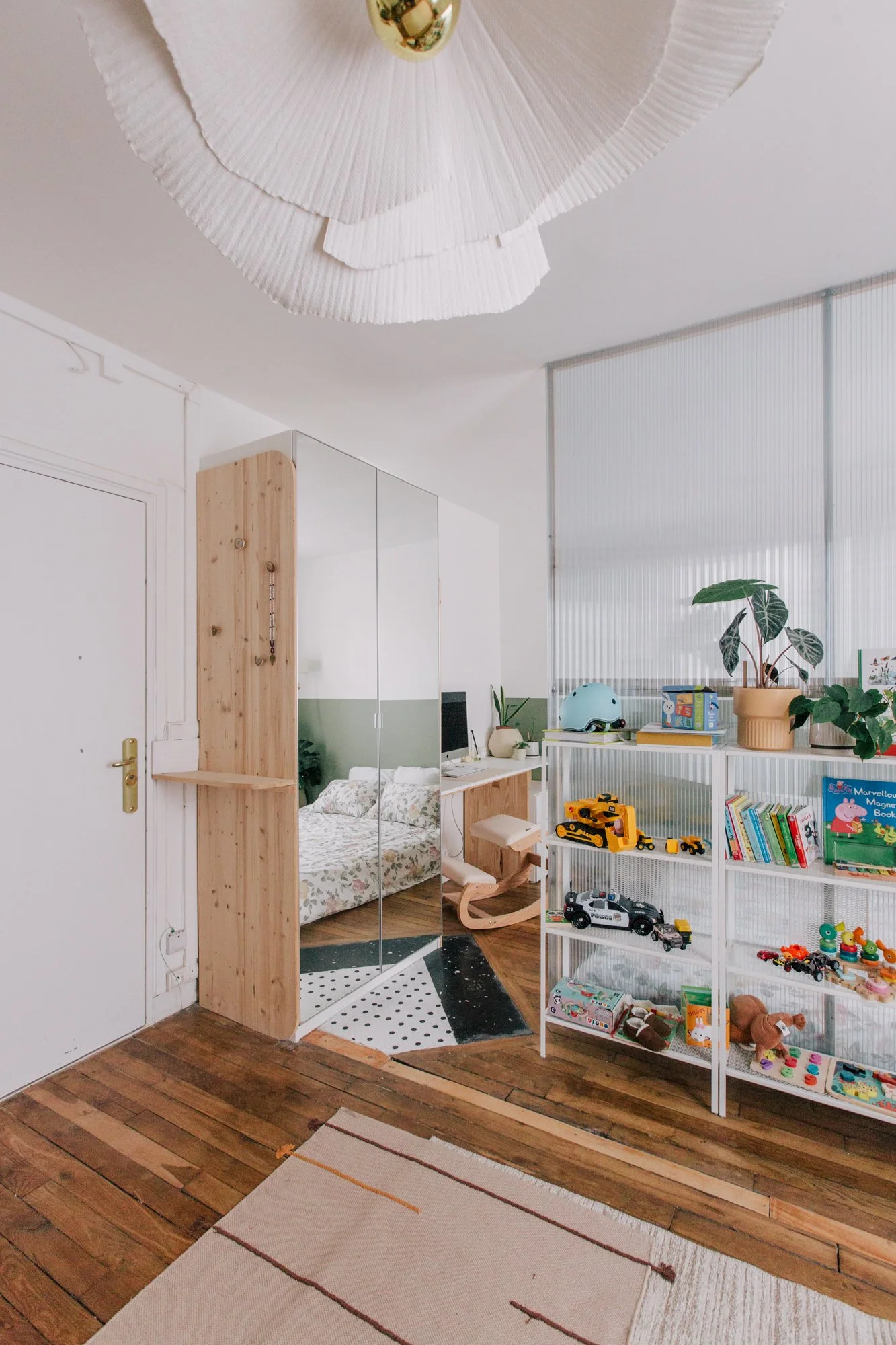 Vue intérieure d'une chambre d'enfant avec un mur blanc, un placard avec miroir, un lit avec une couverture à motifs, une étagère blanche contenant des jouets, livres et divers objets, un parapluie pour bébé, une plante verte, un plafond avec un écla