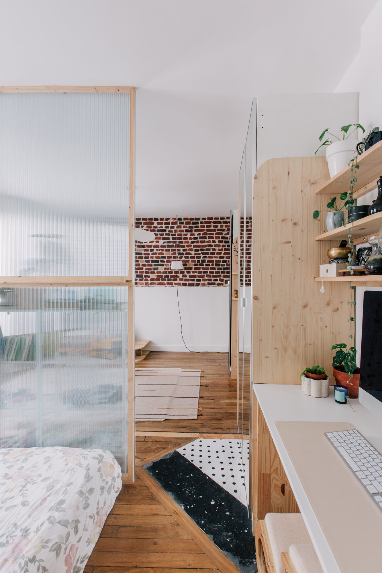 Intérieur d'une pièce avec un lit à gauche, un mur en briques rouges en arrière-plan, une étagère en bois avec des plantes vertes, un bureau blanc avec un clavier, un tapis noir et blanc, et plusieurs éléments de décoration.