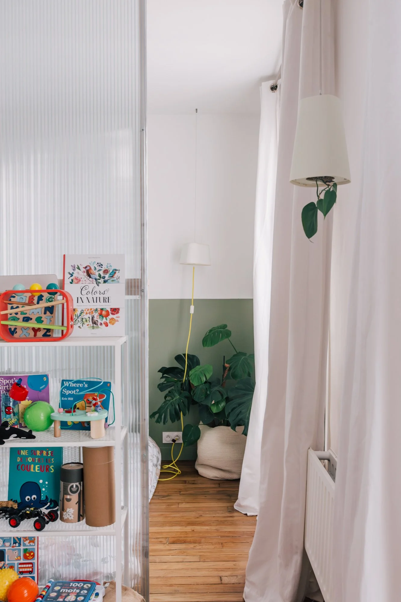 Un coin d'une pièce avec une étagère remplie de jouets pour enfants, des livres et des décorations colorées, un sol en bois, une plante verte à côté d'une lampe, et des rideaux blancs couvrant une fenêtre.
