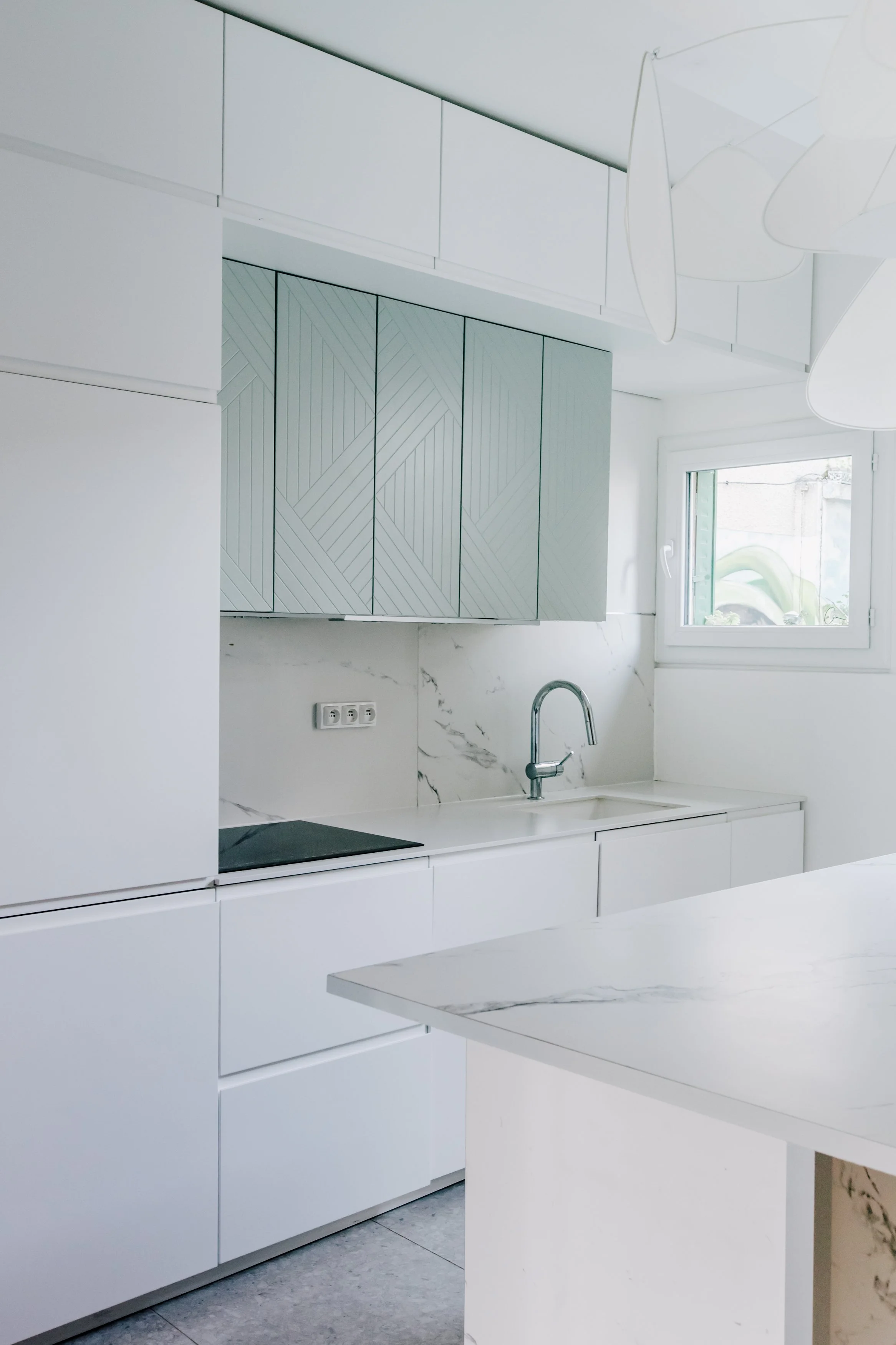 Cuisine moderne blanche avec des armoires lisses, un évier avec robinet, un plan de travail en marbre blanc, et une fenêtre laissant entrer la lumière naturelle.