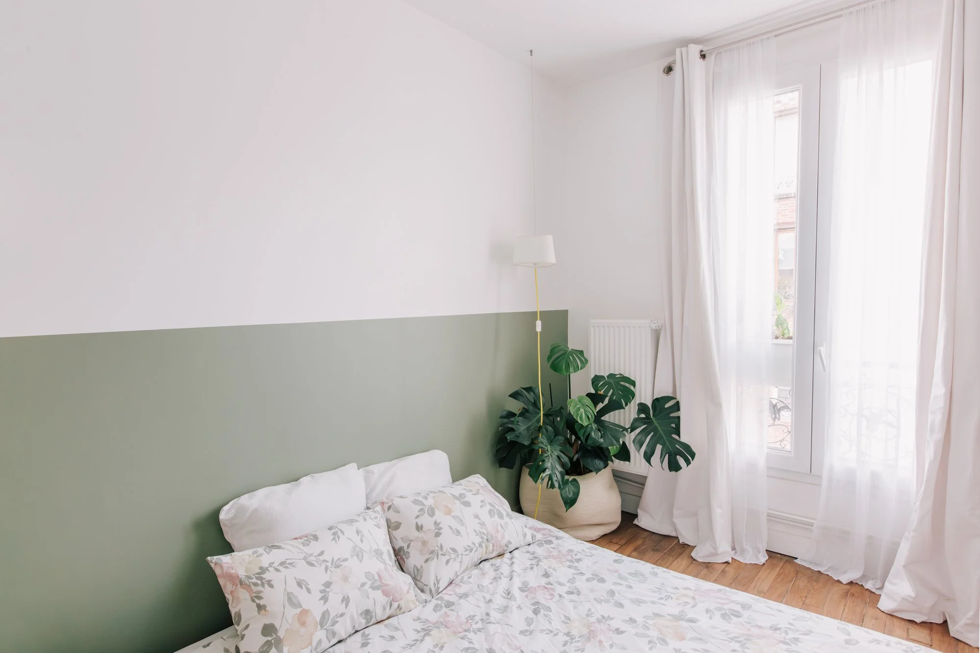 Chambre lumineuse avec lit à les oreillers avec une housse à motif floral, mur peint en deux tons, vert en bas et blanc en haut, grande fenêtre avec rideaux blancs, plante verte près du radiateur, lampe suspendue, parquet en bois.