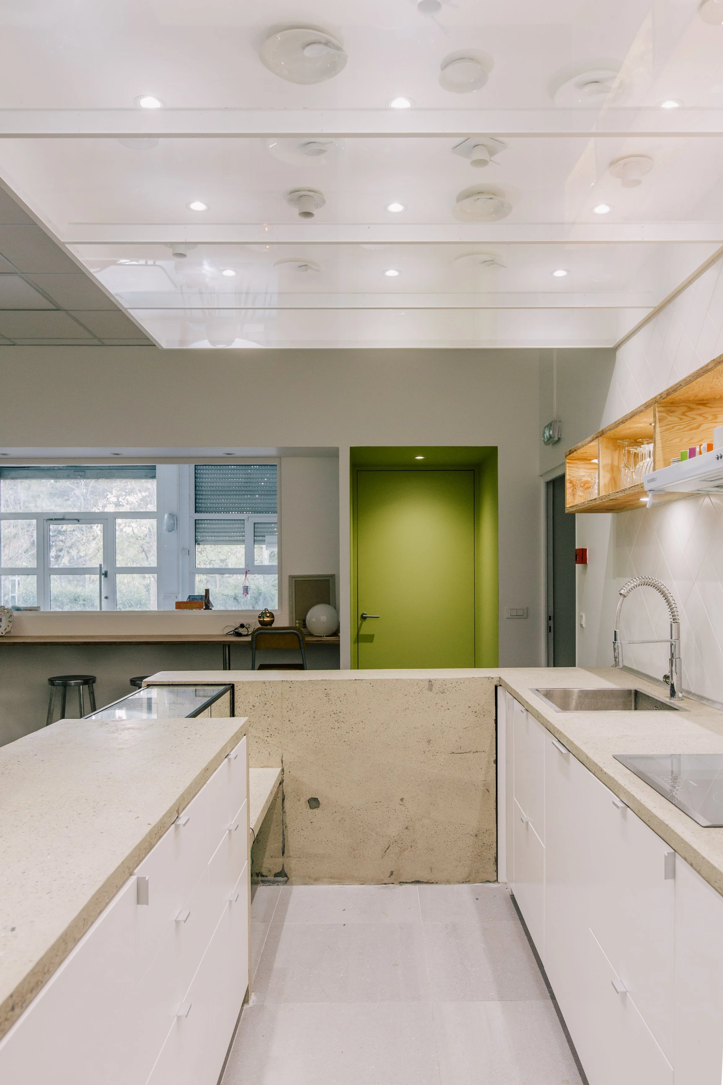 Cuisine moderne avec comptoirs en quartz beige, armoires blanches, évier en acier inoxydable, étagères en bois avec vaisselle et accessoires, mur vert lime, grandes fenêtres laissant entrer la lumière naturelle.