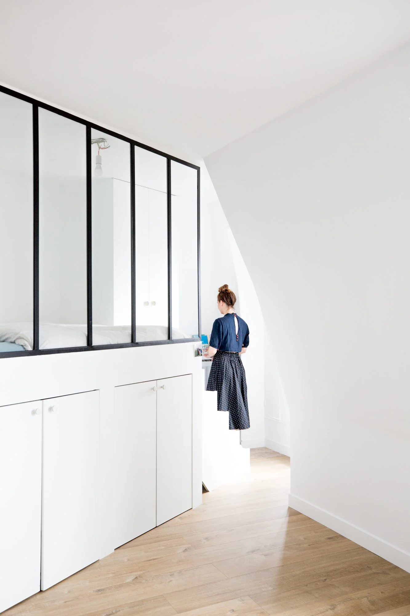 Une femme debout dans une pièce minimaliste blanche, utilisant un ordinateur portable posé sur une armoire blanche. La pièce a un plafond blanc et une grande fenêtre laissant entrer de la lumière naturelle.