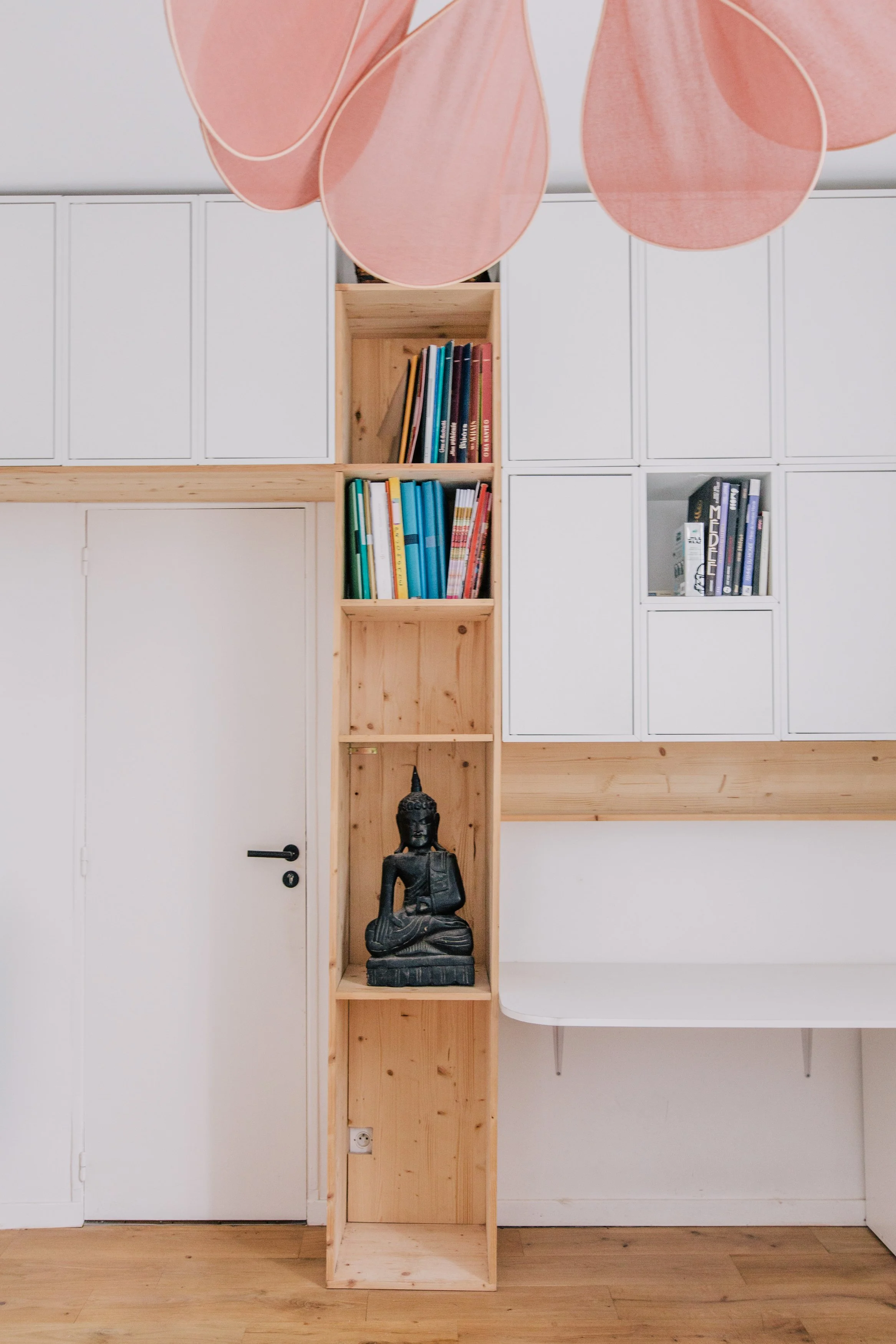 Un mur blanc avec un placard en bois, une étagère en bois avec un livrebot et une statuette de Bouddha noire, et une table blanche en dessous. Un lustre rose suspendu au plafond.