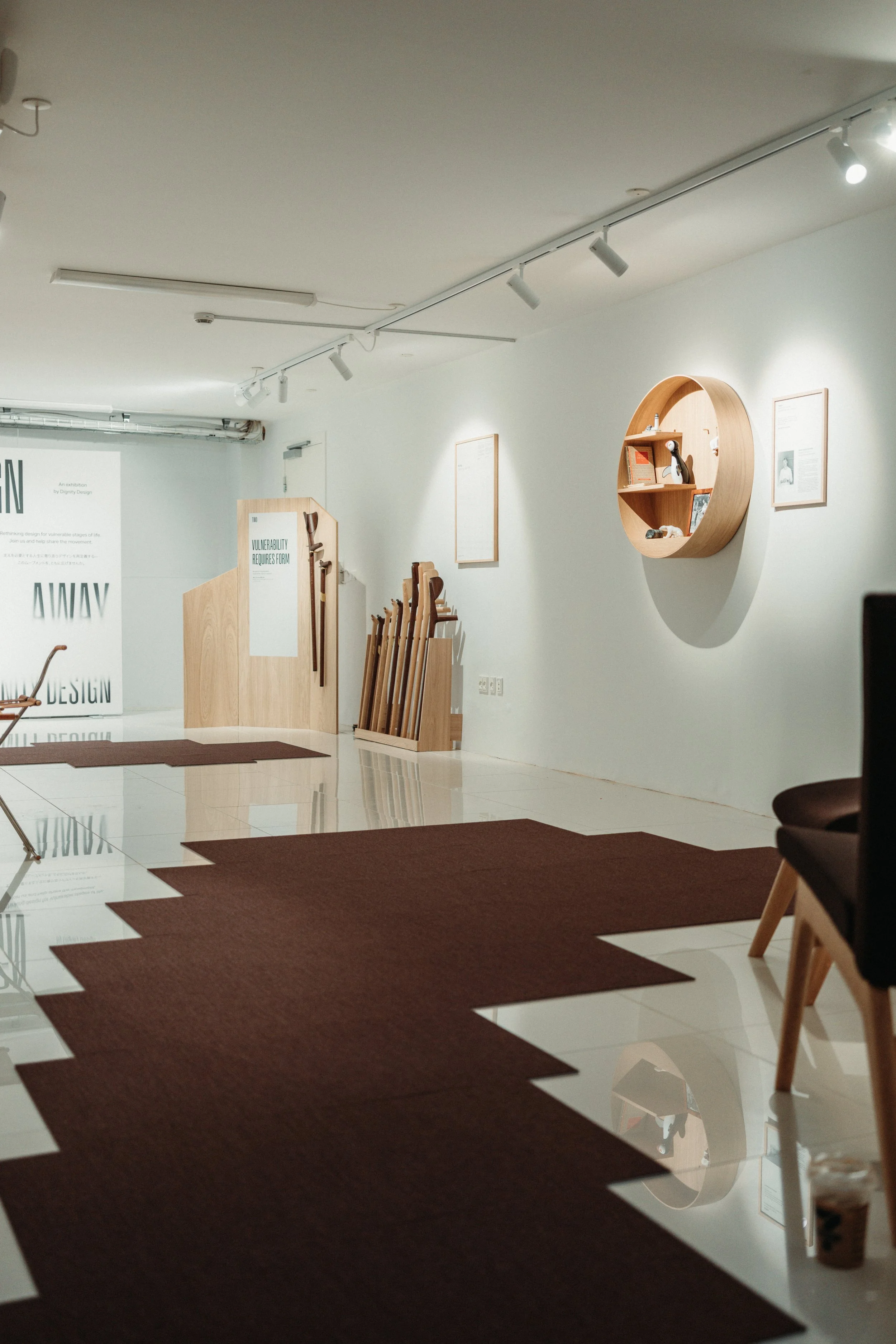 An art gallery interior with white walls, wooden display shelves, chairs, and brown mats on the shiny floor.
