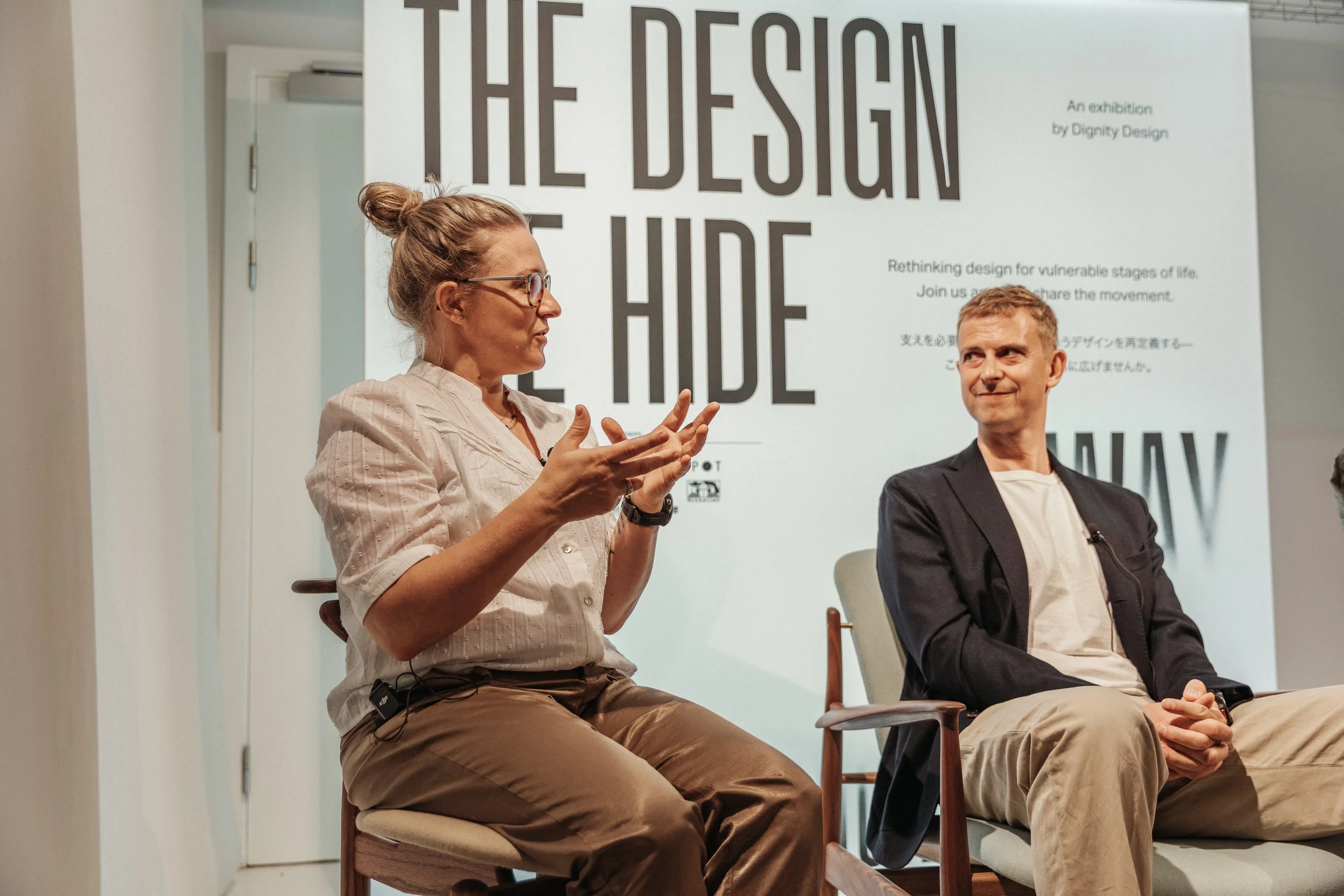 A woman with glasses speaking on a stage during an event titled 'The Design of the Future,' with a man sitting beside her and a large informational poster in the background.