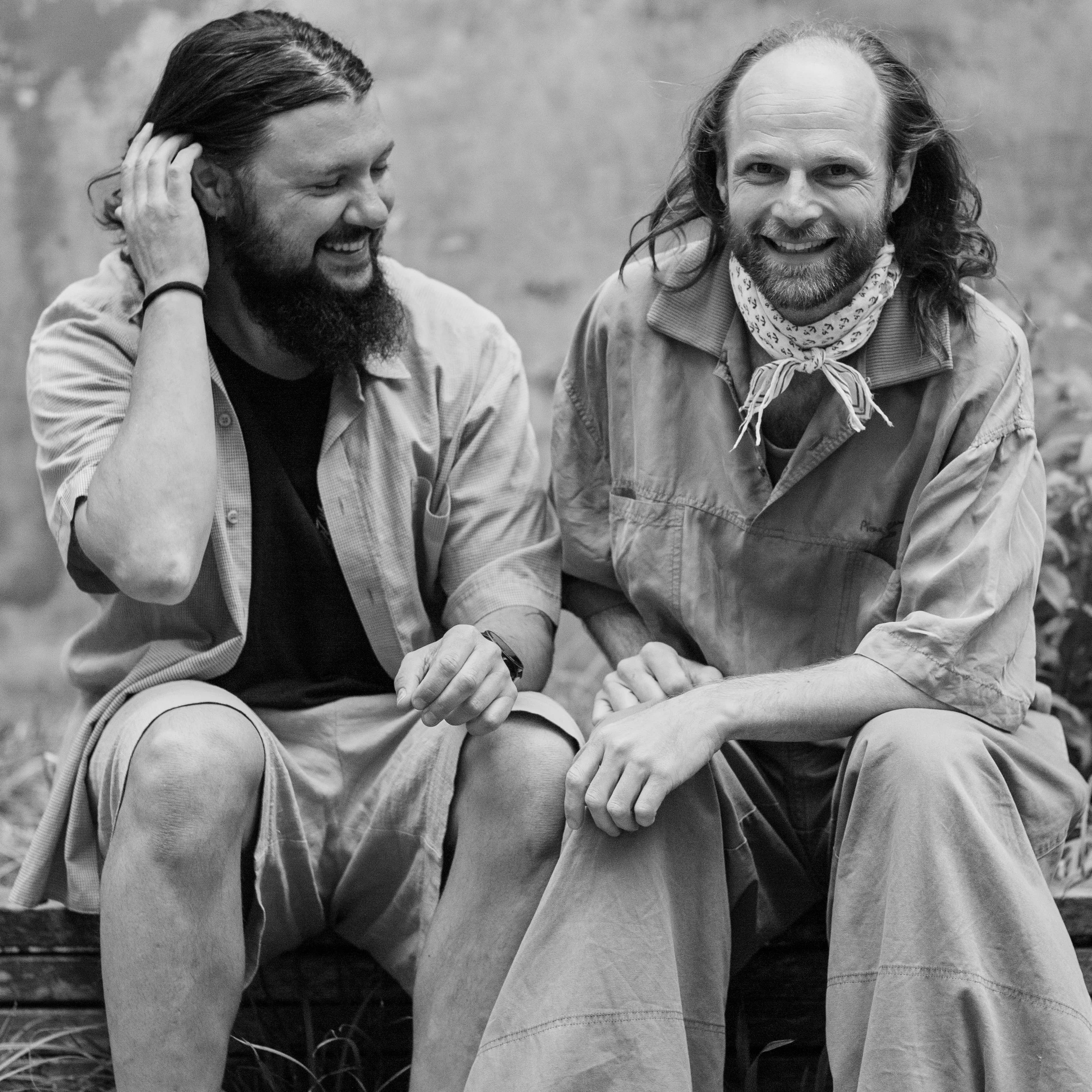 Two men sitting outdoors, smiling and laughing, with one man touching his hair and the other resting his arm on his knee. Both are wearing casual clothing and appear to be enjoying a conversation.