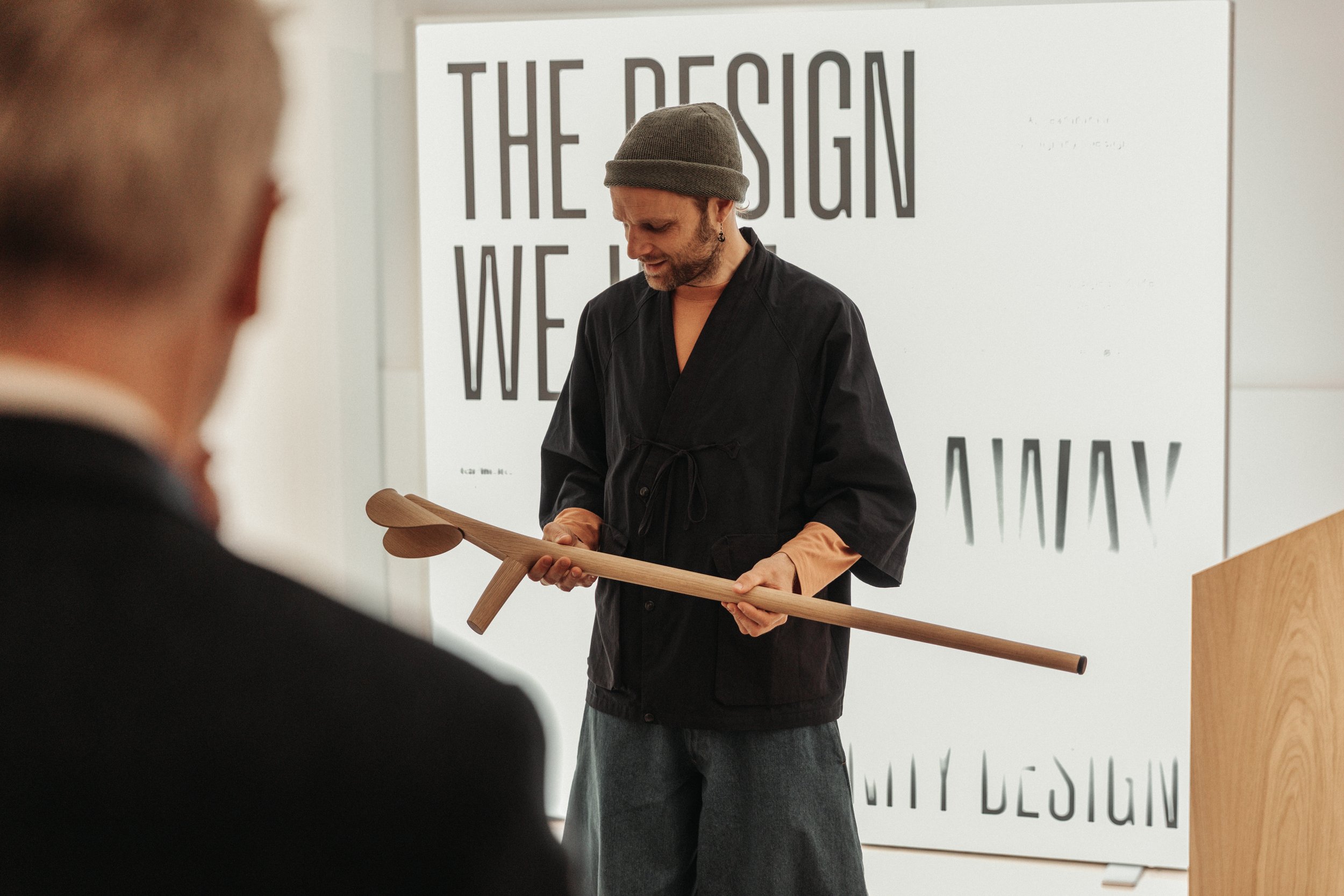 A person wearing a gray beanie and black clothing holding a wooden object in front of a white wall with large text that reads 'THE DESIGN WE LOVE'.